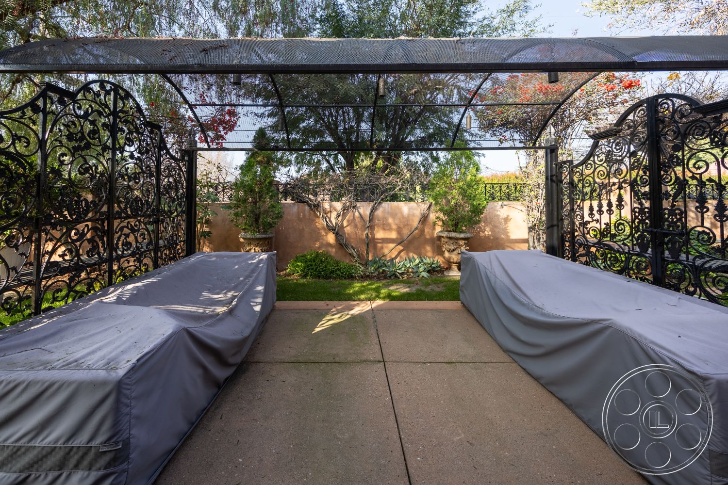 Mansion 145 - smooth concrete pathway, concrete patio flooring, urban outdoor retreat, exterior patio area, covered outdoor seating area, textured stucco wall, garden patio setting, adjacent green lawn, decorative wrought iron gate, landscape with shrubs and trees, covered outdoor space with mesh canopy, garden area with ornamental plants, fenced garden wall, outdoor furniture covers, paved outdoor space