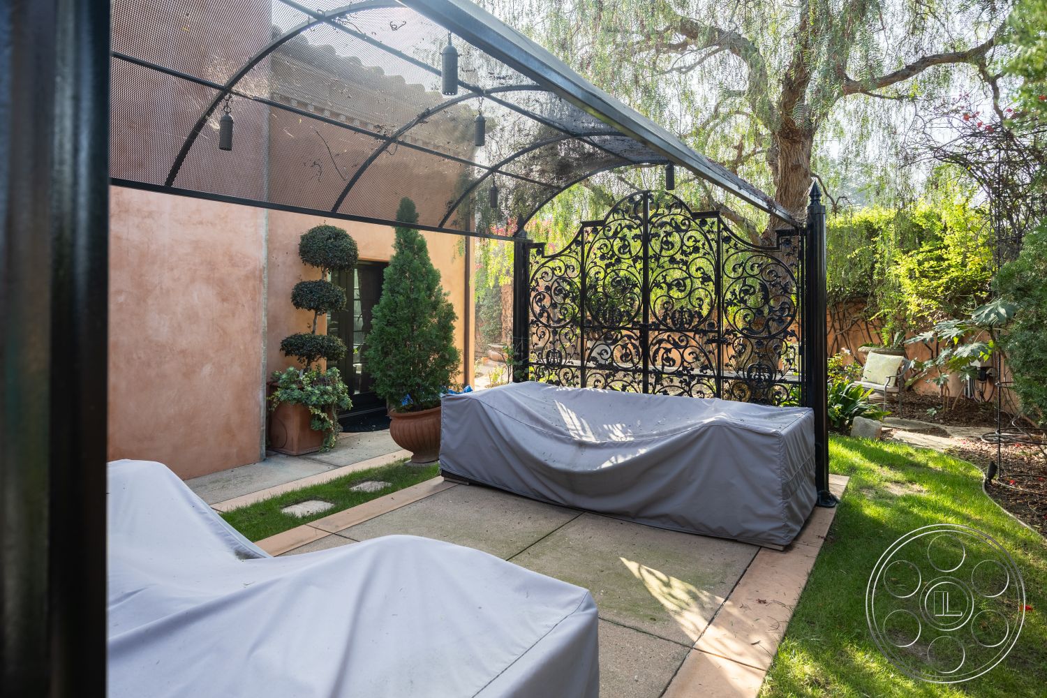 Mansion 145 - al fresco dining area, concrete patio flooring, stucco wall finish, green lawn area, mature trees in background, sunlit garden retreat, exterior courtyard setting, ornate wrought iron trellis, landscape garden with topiaries, paver stone pathways, fabric-covered outdoor furniture, decorative garden accents, covered outdoor seating space