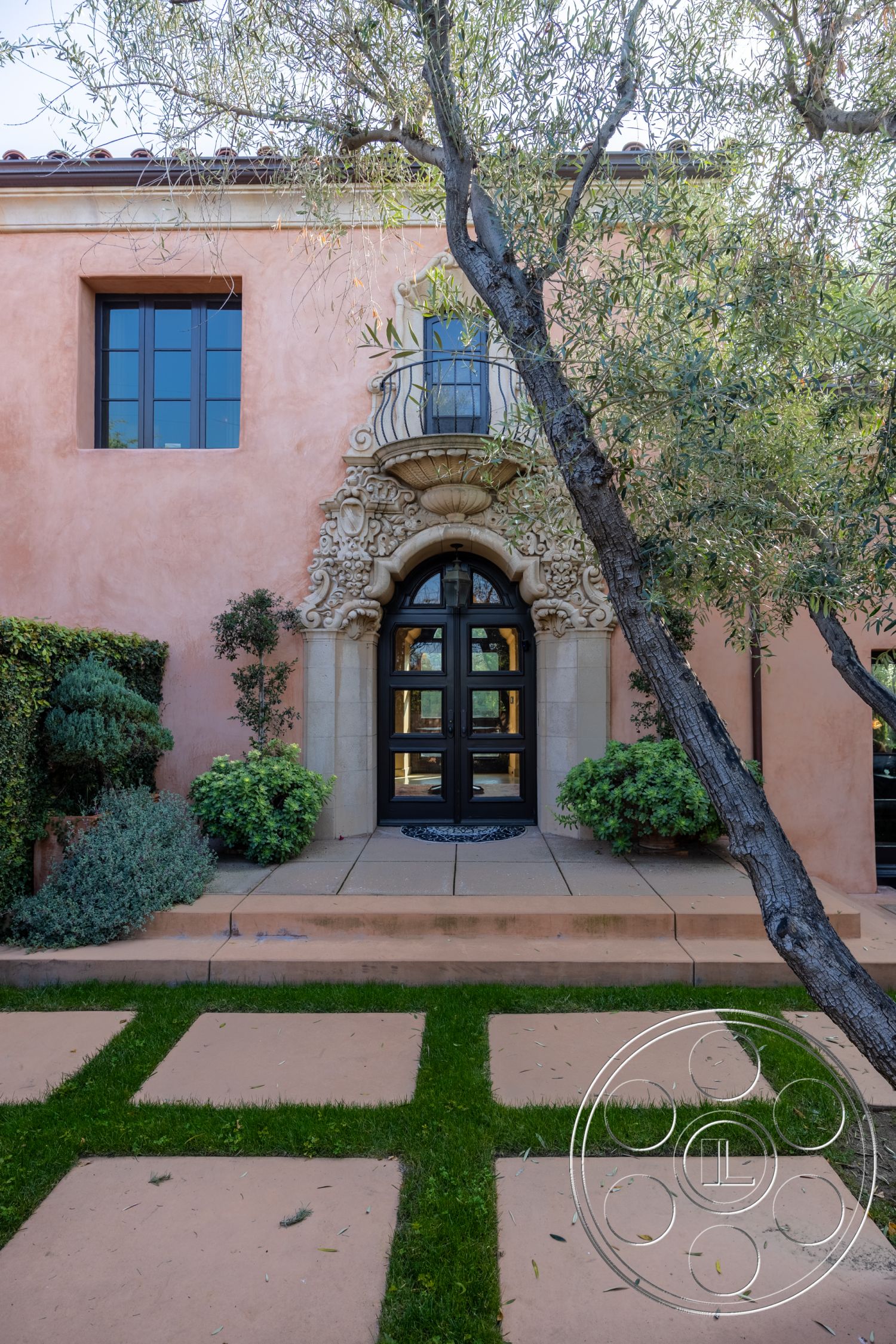 Mansion 145 - exterior, concrete stepping stones, decorative stone planter, stucco wall finish, black wrought iron balcony railing, olive tree landscaping, landscaped garden with shrubs, modern Mediterranean architectural style, ornate stone entrance arch, dark wood double doors, ceramic tile front porch, level lawn