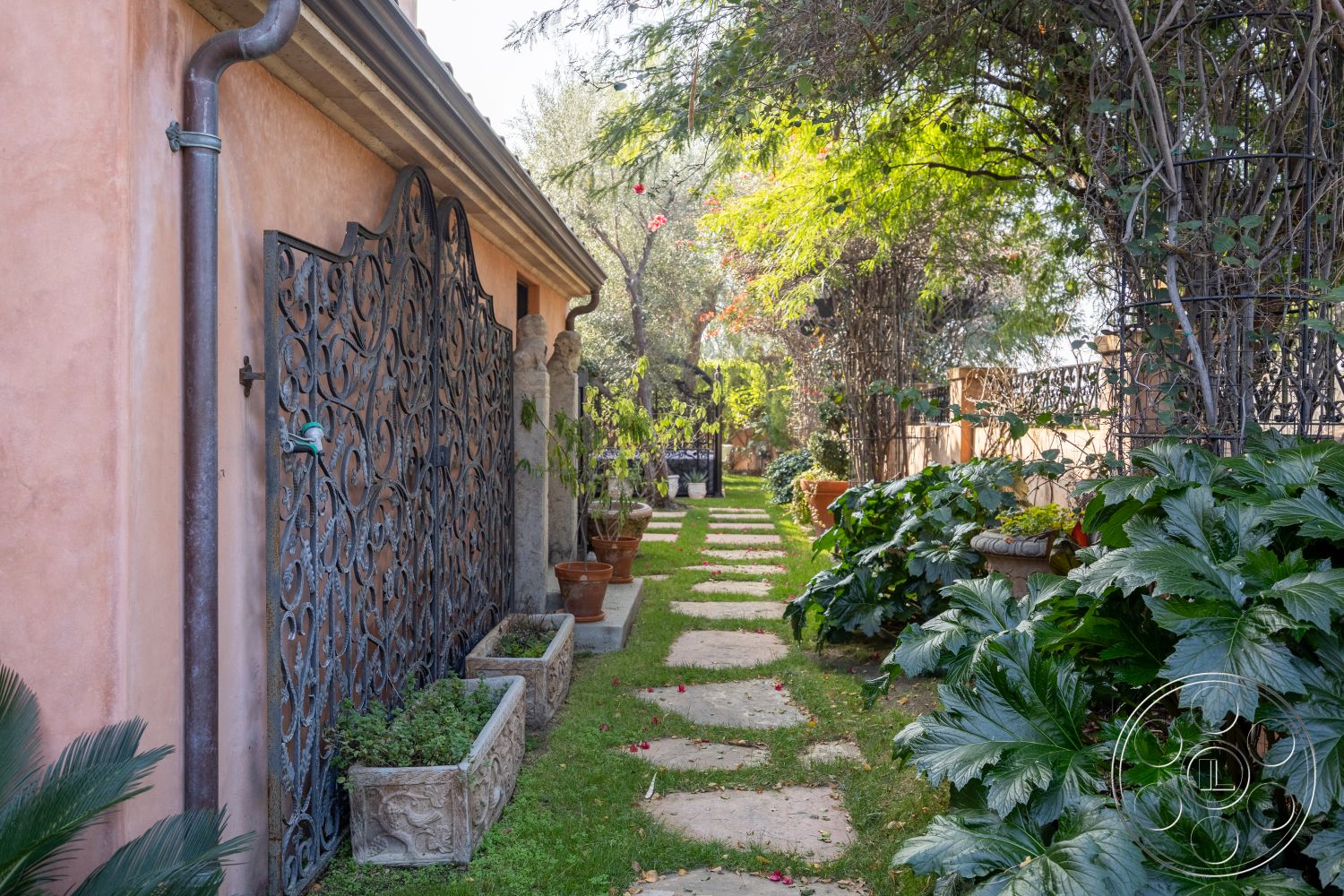 Mansion 145 - exterior, landscaped garden, lush greenery, potted plants, garden setting, pink stucco walls, decorative stone planters, wrought iron gate, curved garden path, stone garden path, urban garden space, ornate wrought iron details, grassy walkway