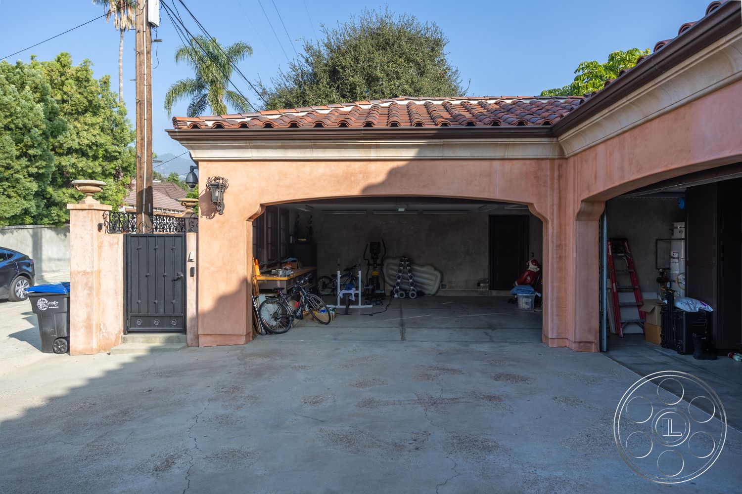 Mansion 145 - exterior, concrete driveway, tile roof, black wrought iron gate, textured concrete floor, modern residential setting, landscaped palm trees, stucco garage walls, arched garage doorway, black outdoor lighting fixture, driveway parking area, rural neighborhood, connected to outdoor space, organized garage storage