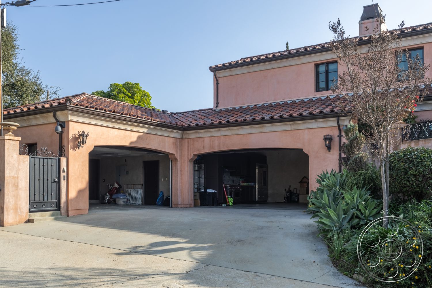 Mansion 145 - landscaped garden, lush greenery, concrete driveway, arched doorways, terracotta tile roof, coastal setting, exterior wall sconces, Spanish style architecture, two-story home, stucco exterior walls, paved patio area, courtyard home, decorative wrought iron details, metal driveway gates, outdoor planter boxes