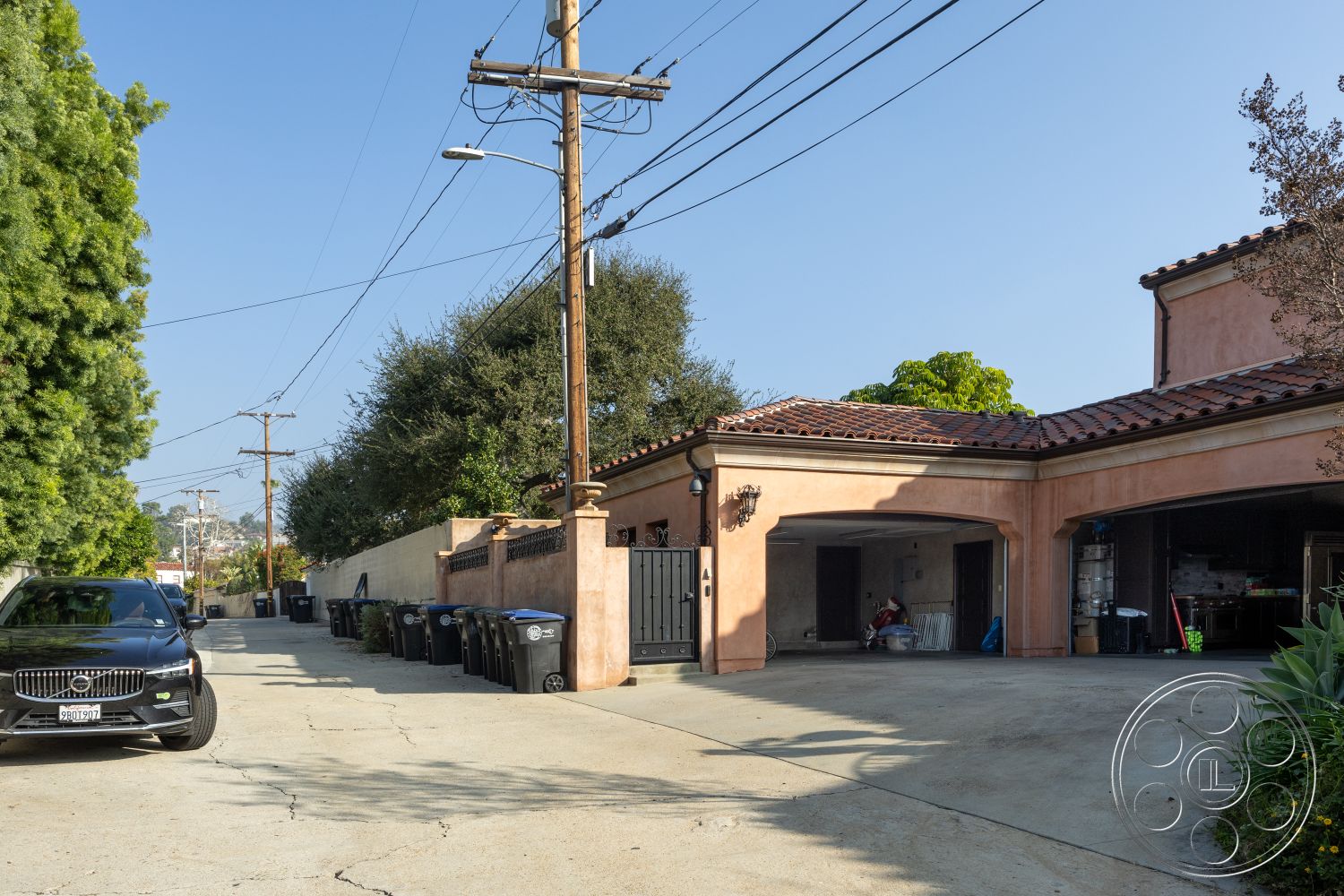 Mansion 145 - stucco facade, tile roof, landscaped garden area, exterior concrete driveway, lush greenery surrounding property, decorative wrought iron gate, alley setting, paved outdoor parking space, several outdoor trash bins, modern black sedan, utility pole with overhead wires