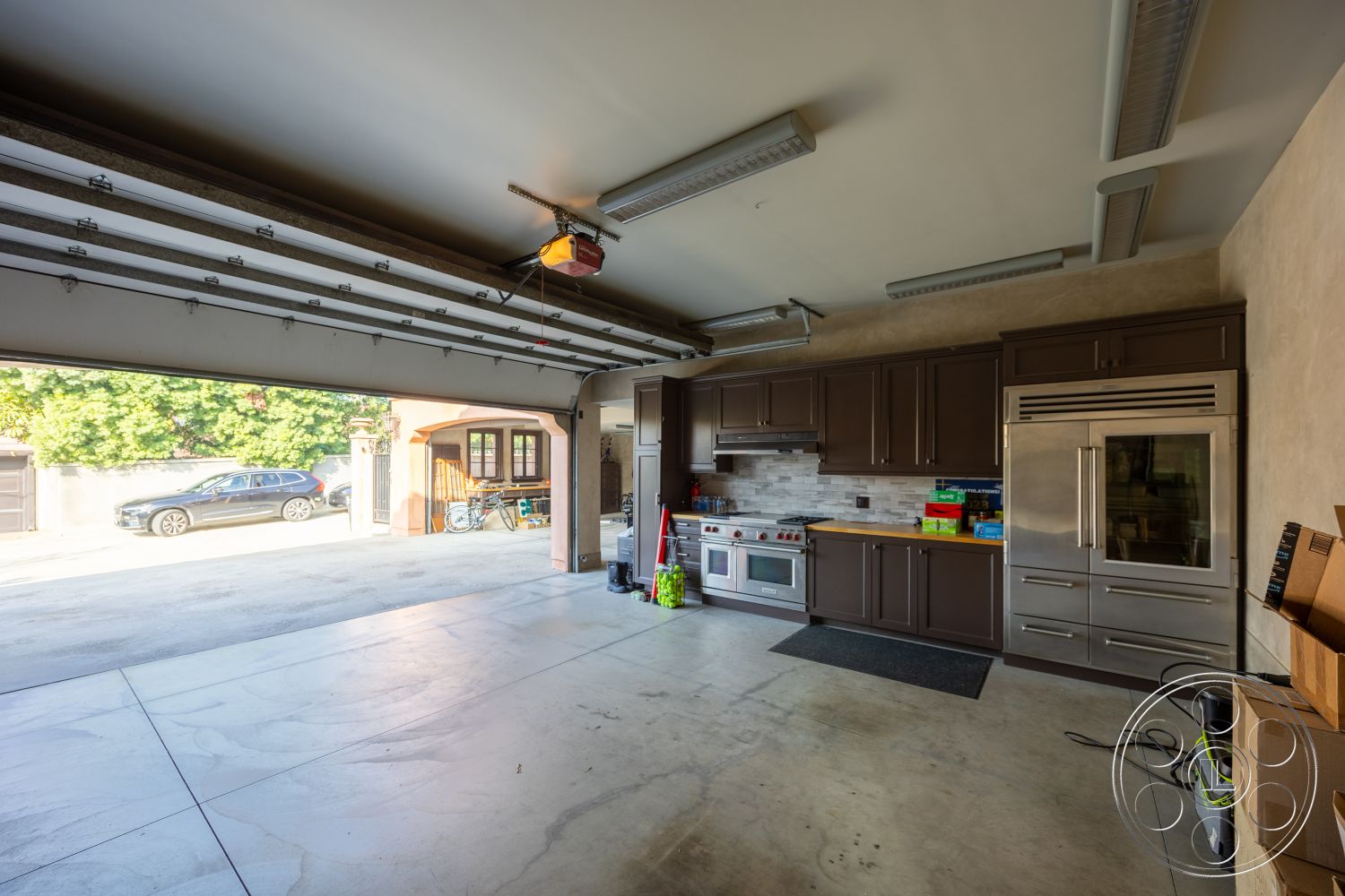 Mansion 145 - dark wood cabinetry, modern lighting fixtures, contemporary design elements, stainless steel refrigerator, stainless steel kitchen appliances, concrete garage floor, industrial-style ceiling, interior garage space, floor-to-ceiling storage cabinets, utility area with appliances, open garage layout, gray tile kitchen backsplash, built-in kitchen area, expanded garage workspace, cabinets with sleek handles, rolling garage door, greenery view from garage, functional and organized, home workshop space