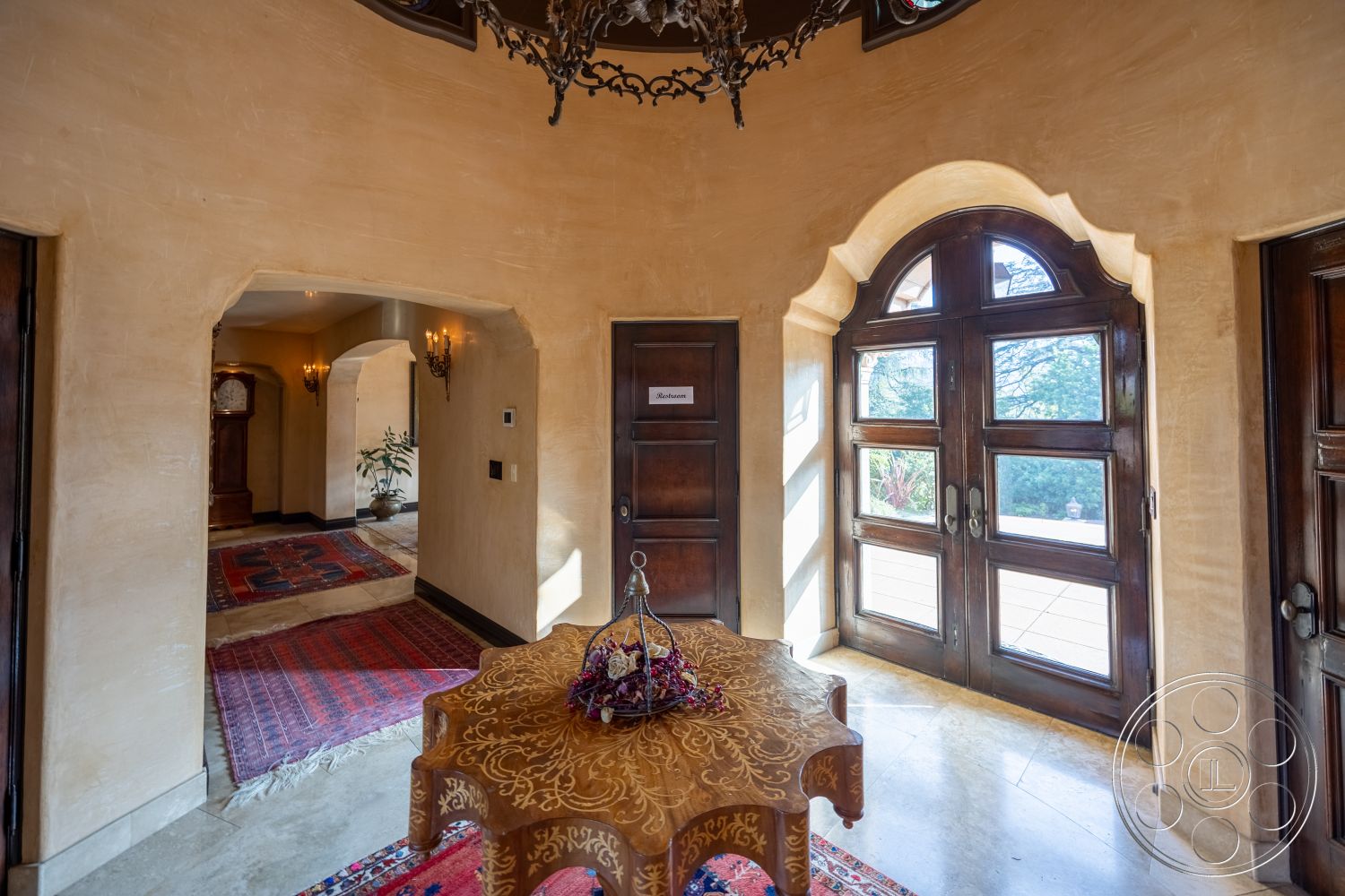 Mansion 145 - indoor greenery, arched doorways, textured plaster walls, dark wood paneling, ornate chandelier, stone tile flooring, traditional area rug, hardwood entryway floors, exterior entrance, interior foyer, warm natural lighting, curved wall structure, wood double doors, decorative accent table, artistic ceiling details