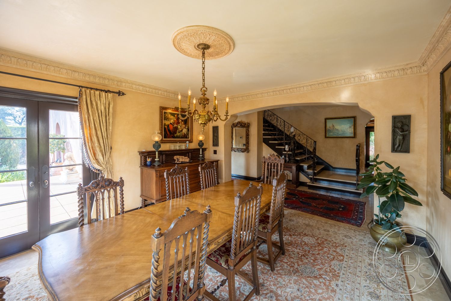 Mansion 145 - arched doorways, textured plaster walls, interior dining room, large windows with garden view, chandelier lighting fixture, traditional Persian area rug, glass French doors, hardwood dining table, potted indoor plant, ornate wooden dining chairs, satin finish crown molding details, decorative wall paintings, dark wood stair railing, curved stairs, wooden sideboard cabinet, elegant silk drapery