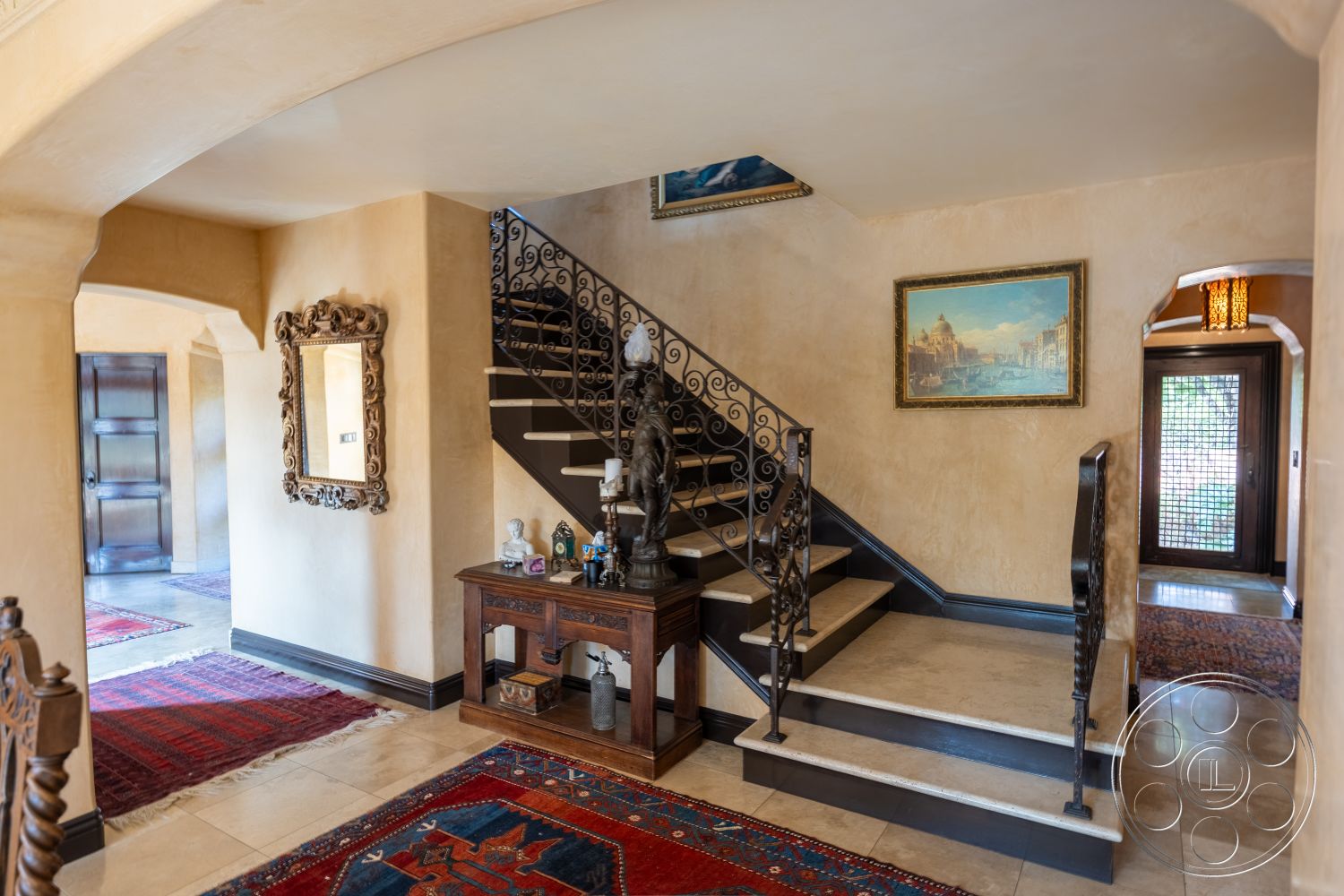 Mansion 145 - textured plaster walls interior, ornate wrought iron stair railings interior, polished stone steps interior, dark wood entry door exterior, traditional Persian area rug interior, framed landscape painting wall art interior, archway entrance interior, recessed lighting ceiling fixtures interior, glazed ceramic tile flooring interior, decorative wood console table interior, neutral beige color palette interior, intricate mirror frame interior, multicolored patterned area rug interior, high ceiling with crown molding interior, vaulted entryway ceiling interior, open staircase design interior, classic architectural details interior