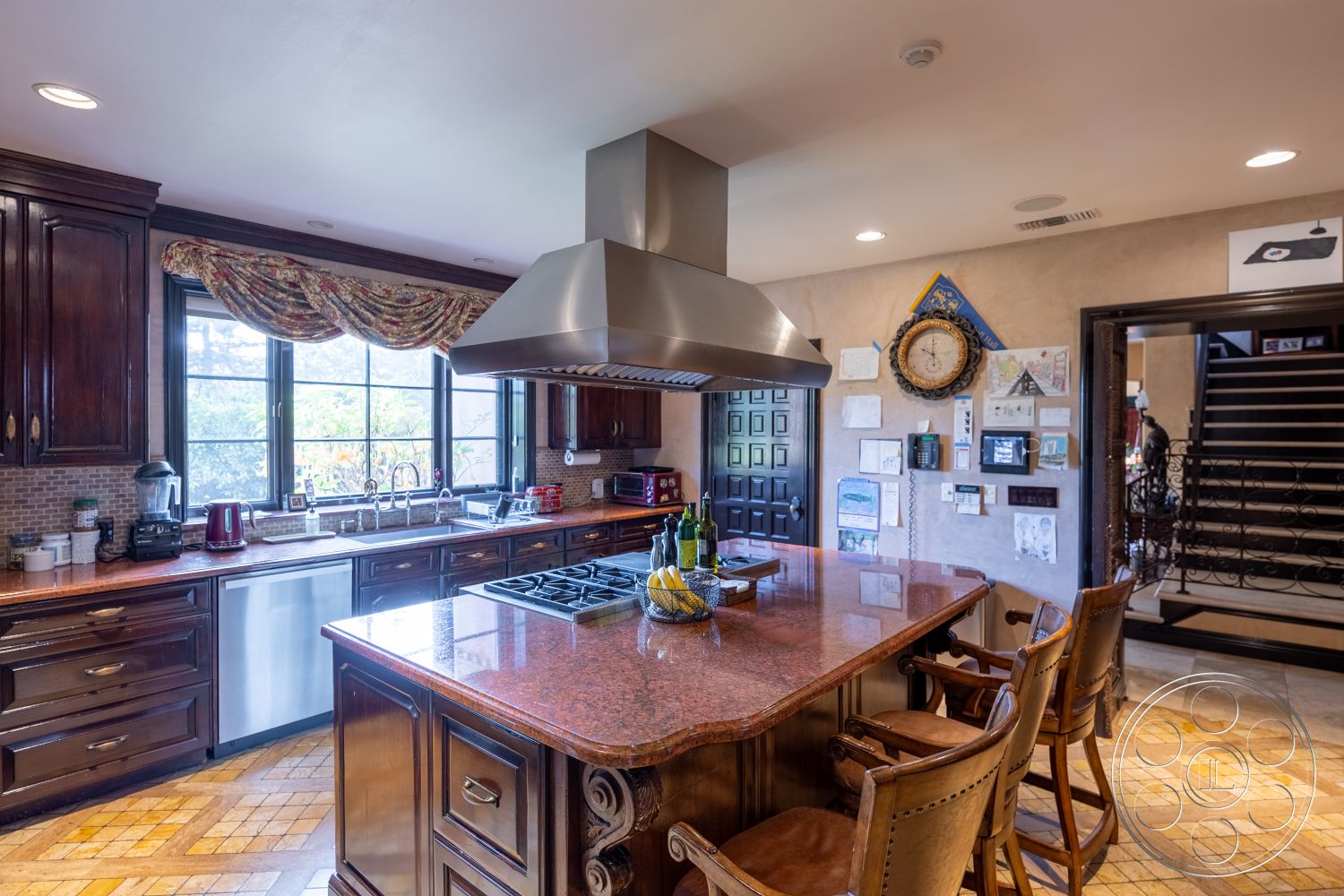 Mansion 145 - interior kitchen with cherry wood cabinetry, granite kitchen island countertop with decorative edge, stainless steel vent hood above gas cooktop, under-cabinet lighting on wood paneling, black framed windows with garden view, mosaic tile backsplash in warm tones, decorative crown molding details on cabinetry, wood parquet flooring with inlaid design, kitchen with open layout to dining area, comfortable bar stools with leather upholstery, traditional kitchen clock on feature wall, wall-mounted display of children's artwork, rich wood panel doors leading to staircase, elegant window treatments with patterned fabric, warm ambient lighting from recessed fixtures