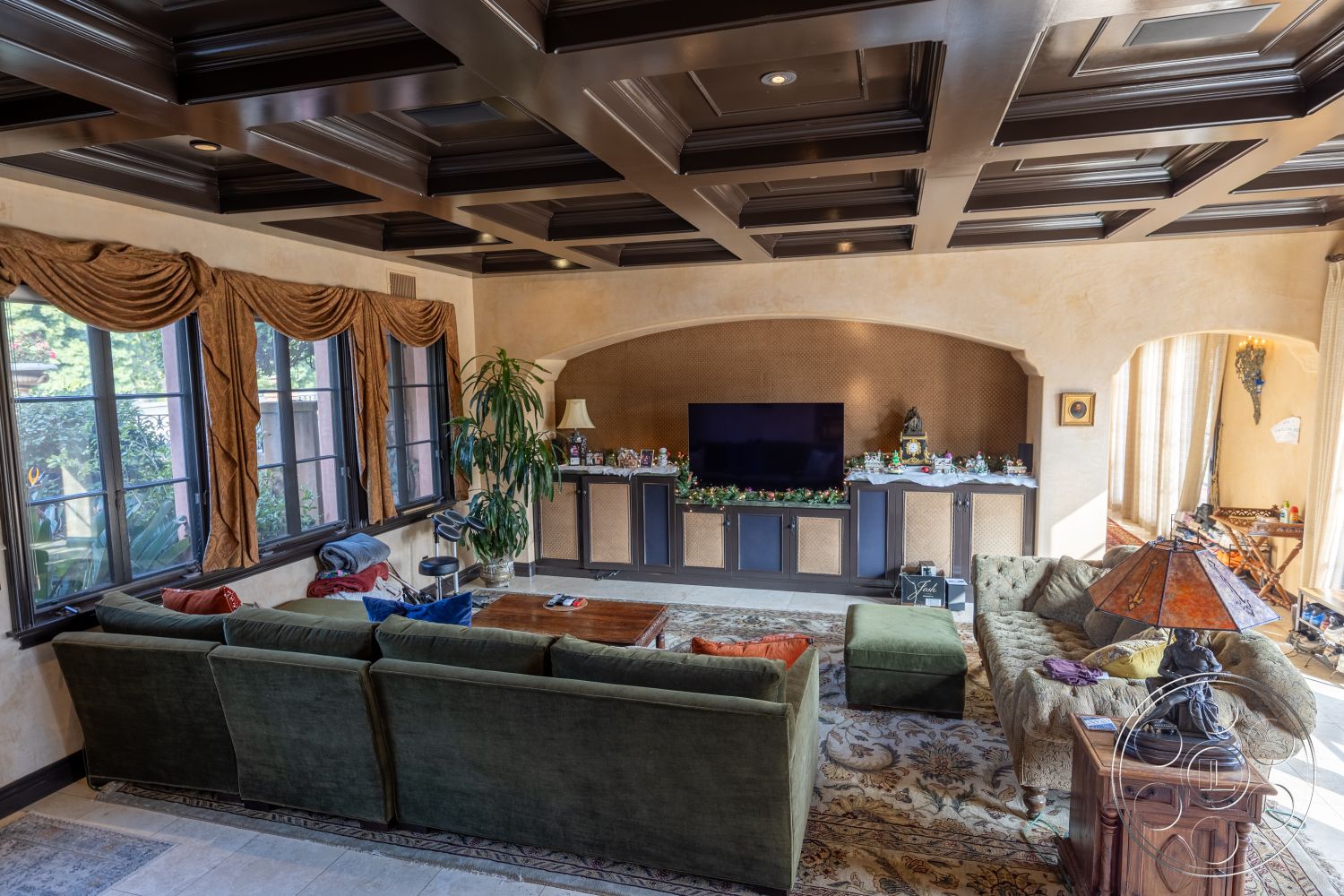 Mansion 145 - hardwood flooring, warm color palette, dark wood furniture, traditional style, built-in entertainment center, decorative accessories, textured wall finishes, area rug with intricate patterns, spacious seating area, luxurious living space, plastered interior walls, dark wood paneling accents, architectural moldings, large double-hung windows, rectangular living room layout, houseplant decor, modern television setup, coffered dark wood ceiling, tapestry drapes, artistic table lamps