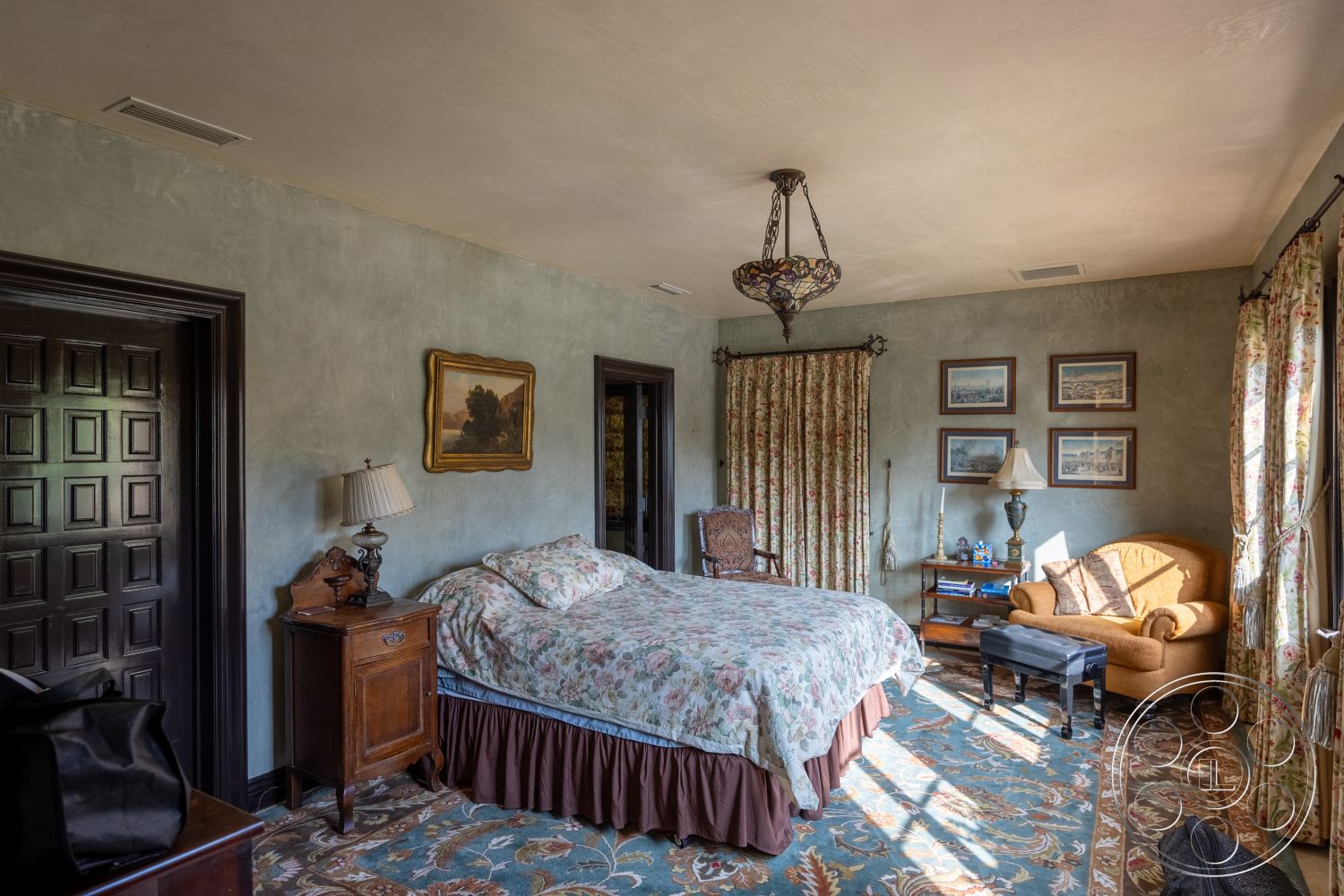 Mansion 145 - hardwood bedroom floors interior, dark wood paneling interior, textured plaster walls interior, floral patterned area rug interior, classic chandelier light fixture interior, wooden framed wall art interior, elegant curtains with floral design interior, traditional wooden bedside table interior, upholstered armchair with vintage aesthetic interior, glass lamp with fabric shade interior, cozy quilted bedspread interior, antique wood chair interior, detailed door design interior, decorative wall molding interior, vintage travel-themed artwork interior