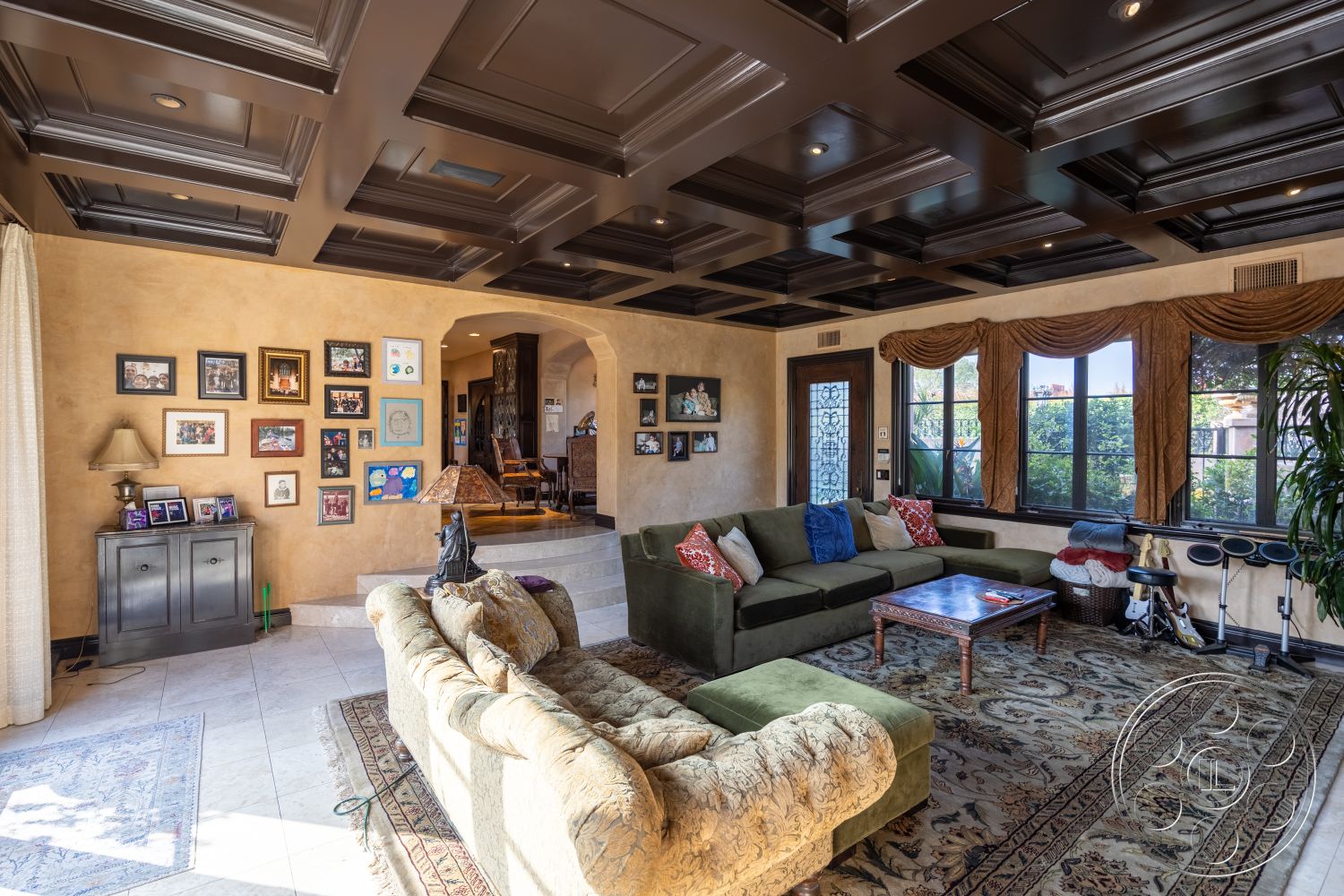 Mansion 145 - open layout connecting to dining area, exterior house with Mediterranean architectural style, interior living room with coffered ceiling, decorative ceiling with dark wood paneling, exposed textured plaster walls, tile flooring with intricate patterns, plush sectional sofa with green upholstery, ornate area rug with traditional motifs, built-in cabinetry with black finish, large windows with decorative drapery, family photo gallery wall, accent lighting integrated into ceiling, custom chandelier hanging from ceiling, arched doorways leading to adjacent spaces, wooden coffee table with rich finish, decorative wall accents and frames, plant accents for natural decor, light-filtering curtains throughout room