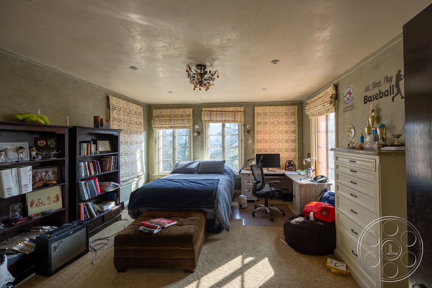 Mansion 145 - neutral color palette interior, textured plaster walls interior, natural light from windows interior, carpet bedroom flooring interior, wooden bookshelf with decorative items interior, large windows with patterned shades interior, queen-size bed with blue bedding interior, desk area with computer and office chair interior, cozy ottoman with colorful accessories interior, ambient ceiling lighting fixture interior, baseball-themed wall art interior, trophy display on dresser interior, functional home office space interior