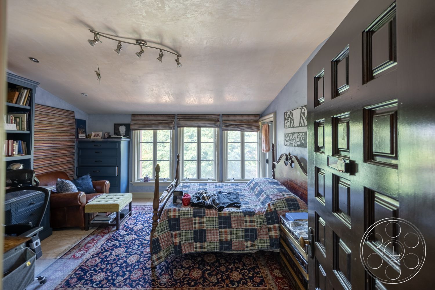 Mansion 145 - textured plaster walls, exposed wood ceiling beams, built-in bookshelf with decorative items, blue painted accent wall, interior bedroom with checkered bedspread, hardwood floor at the entrance, plush area rug in vintage design, large windows with wooden blinds, dark wood entry door with panel details, cozy reading nook with leather armchair, intricate patterned window treatments, modern track lighting fixtures, comfortable footstool in front of chair, neutral color palette with splashes of color, decorative wall art above bed, small bedside table with decorative lamp, sliding window view of natural surroundings