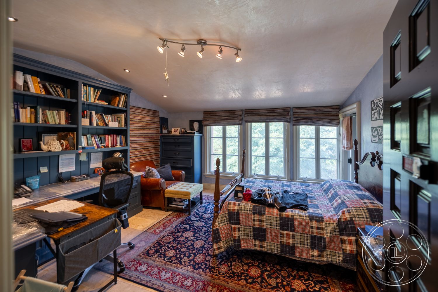 Mansion 145 - neutral color palette, decorative area rug, wooden dresser, textured plaster walls, exposed wood ceiling beams, hardwood bedroom floors, large double-hung windows, interior soft lighting, blue built-in bookshelves, plaid bedspread on queen bed, metal track lighting, cozy reading chair, dark door frame, functional home office space, exterior forest view