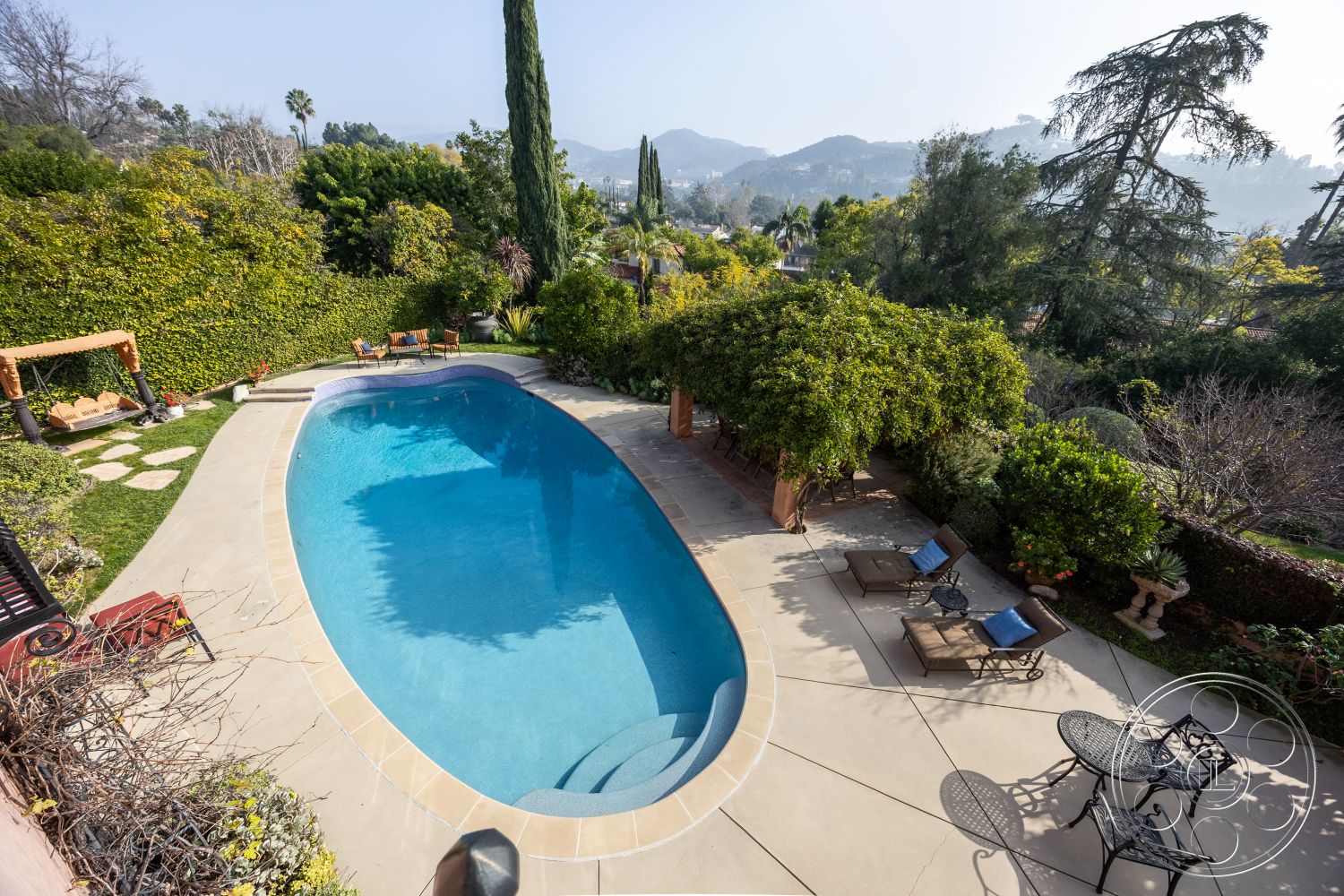 Mansion 145 - landscaped garden setting, stone patio flooring, sunny outdoor environment, lush greenery landscaping, concrete steps leading to patio, mountain view backdrop, private backyard oasis, poolside relaxation area, oval swimming pool exterior, concrete pool deck surrounding pool, outdoor lounge area with chaise lounges, wooden swing set accent, outdoor dining set with metal chairs