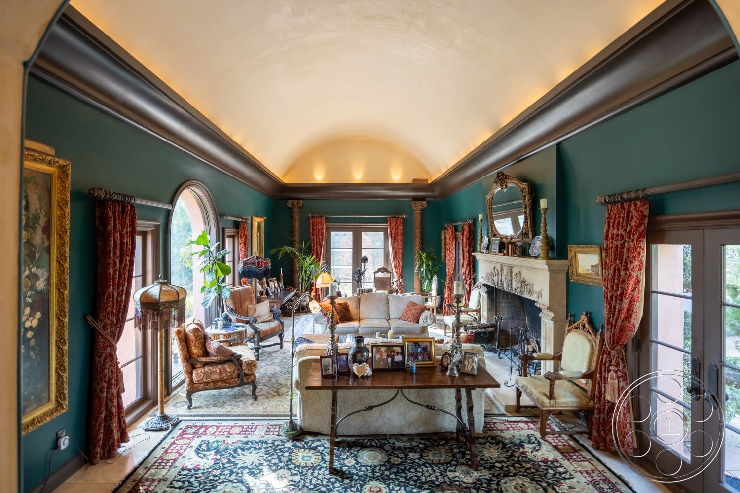 Mansion 145 - hardwood flooring, dark wood furniture, arched doorways, textured plaster walls, interior living room, potted indoor plants, soft ambient ceiling lighting, vaulted ceiling with crown molding, cream-colored marble fireplace surround, richly patterned area rug, elegant window treatments with brocade fabric, decorative wall art with vintage frames, pillowed upholstered seating, traditional lounge chairs, courtyard view through large windows, neutral-toned wall paint, ornate mirror above fireplace, wooden coffee table with decorative items