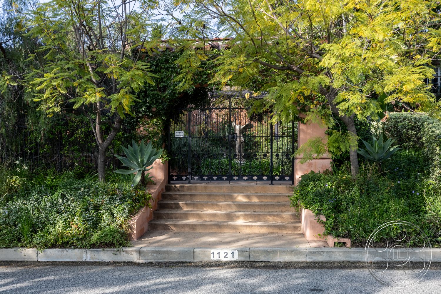 Mansion 145 - clear blue sky, suburban environment, low-maintenance xeriscaping, textured plaster wall, lush garden landscaping, concrete steps with stone border, planted shrubbery, decorative wrought iron gate, exterior gate entrance, Mature trees providing shade, succulent plants in garden beds, curved plant beds, weathered stone pathway, tropical foliage surrounding entrance, driveway with residential curb appeal