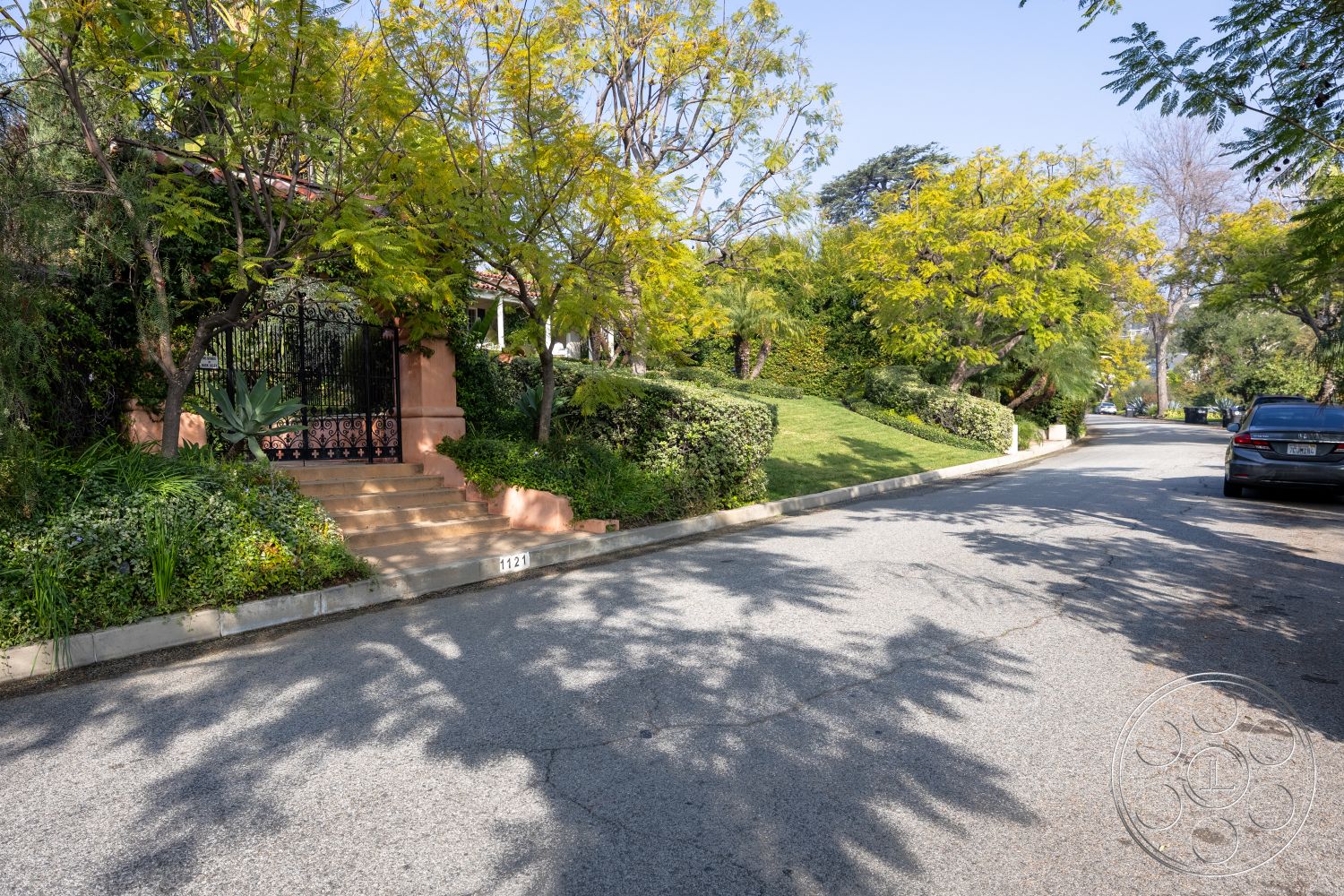 Mansion 145 - suburban setting, open lawn area, paved walkway, curved sidewalk, vibrant flower beds, ornamental trees, lush green landscaping, wrought iron gate, exterior garden area, natural stone garden edging, stepped concrete entrance steps, asphalt residential street, textured concrete driveway border, decorative stucco wall, shade-providing trees