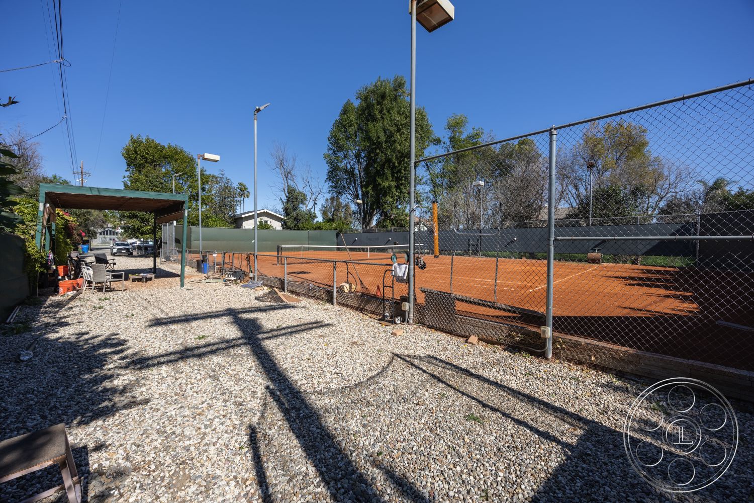 Clay Court - exterior, outdoor seating, decorative planters, hardscape elements, residential backyard, low-maintenance landscape, outdoor lighting fixtures, landscaped garden area, gravel patio flooring, natural sunlight exposure, wooden pergola structure, metal chain-link fencing, clay tennis court surface, fenced tennis court, urban tennis facility setting, concrete support posts