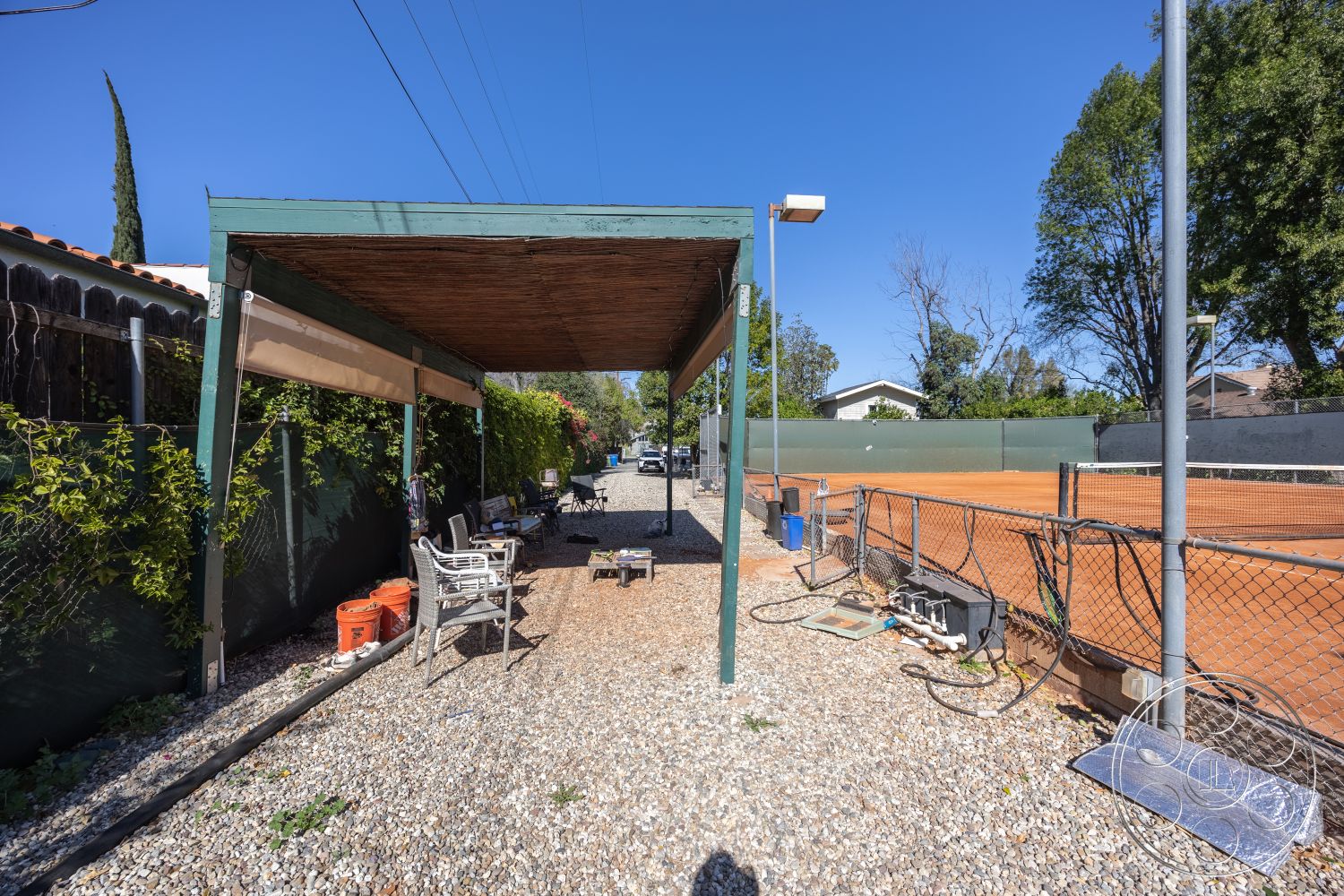 Clay Court - paved driveway, outdoor lighting fixtures, suburban backyard setting, gravel outdoor flooring, exterior pergola structure, fenced tennis area, wood slatted roof covering, green painted steel frame, surrounding tennis court, seating area with lawn chairs, landscaped garden with ivy, wood beam support columns, tree-lined environment