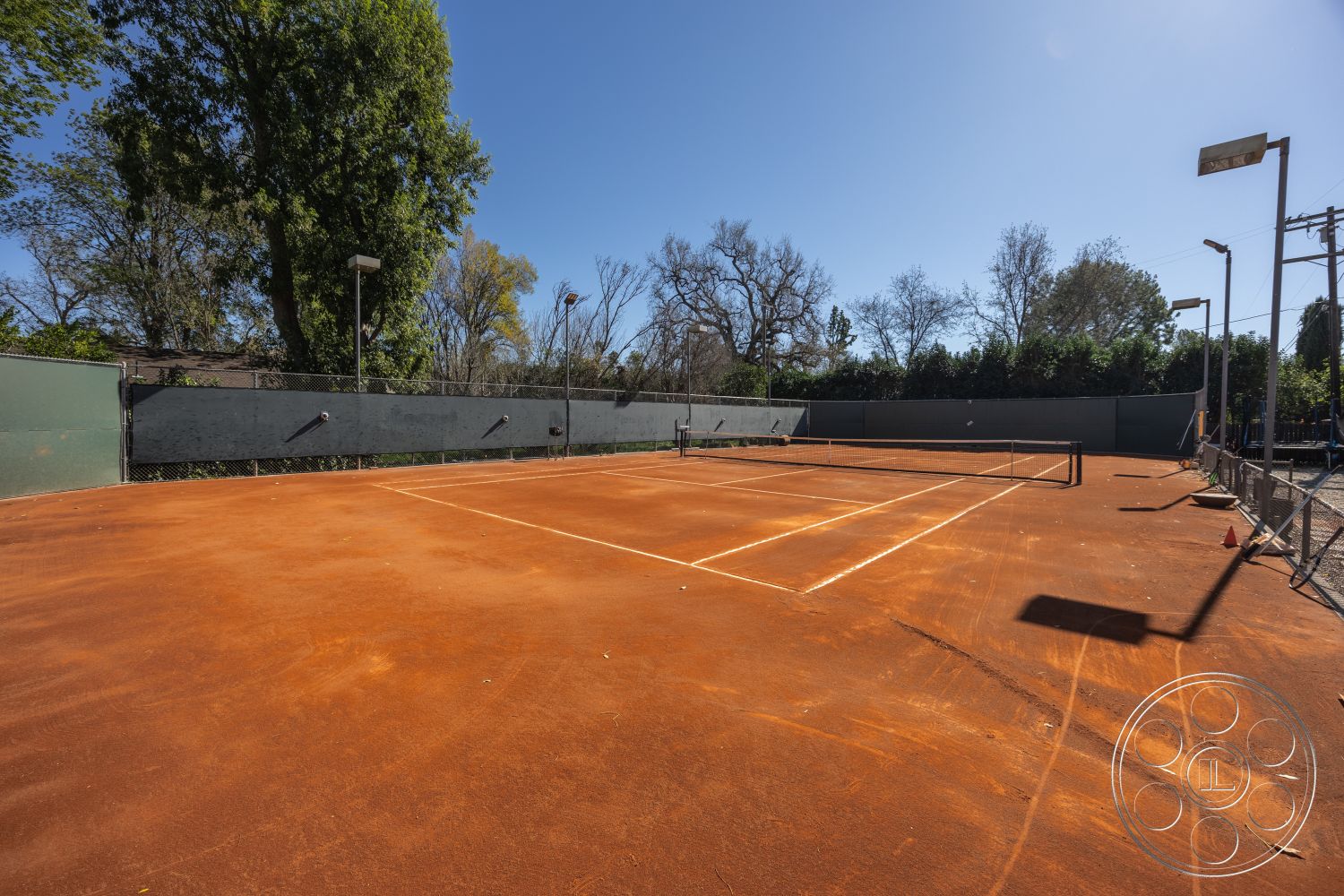Clay Court - outdoor recreational space, clay court, tennis court surface with clay texture, orange clay tennis court, exterior sports court, fenced tennis area with green mesh fencing, surrounding greenery with trees, exterior lighting poles for night play, well-maintained sports facility, hardscape tennis court construction, open sky view above tennis court
