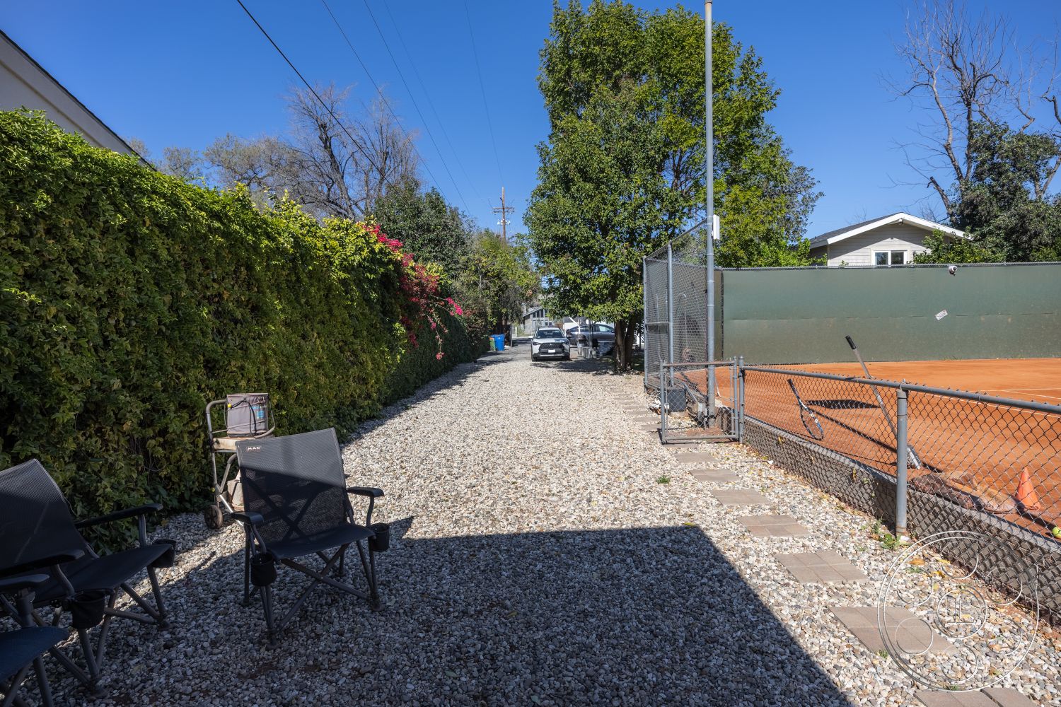 Clay Court - exterior view, outdoor seating area, gravel pathway, paved walkway, urban garden, residential backyard, fenced yard, suburban environment, sunny outdoor setting, decorative patio stones, lush hedge privacy wall, tennis court fencing, landscape foliage, tennis court adjacent, natural shade conditions, stone pathway border