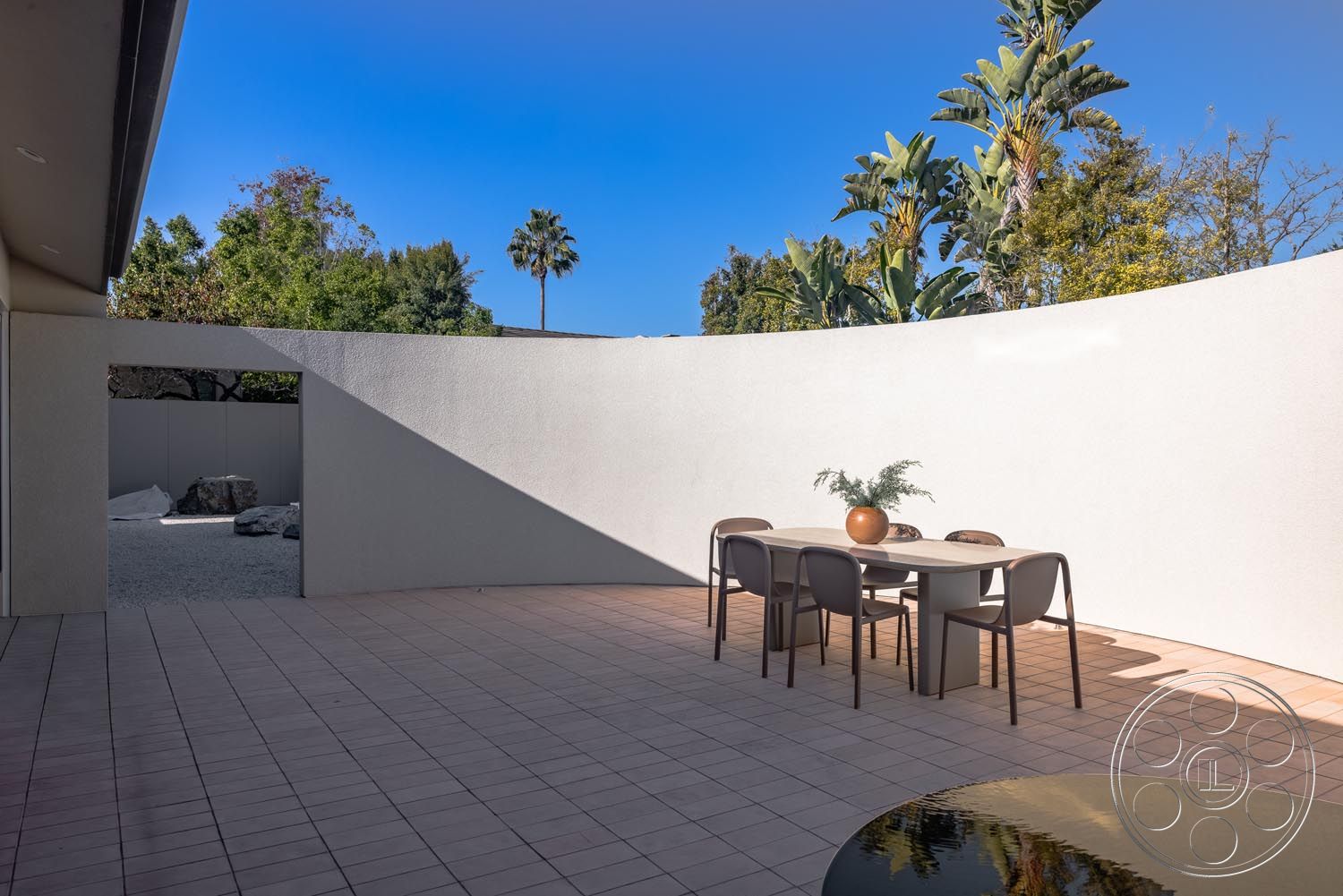 Modern 521 - natural stone accents, textured wall finish, contemporary design elements, minimalist architecture, reflective water feature, surrounding palm trees, open-air dining area, desert environment setting, exterior patio with paver flooring, curved white stucco wall, outdoor dining table with modern chairs, landscaped garden with green foliage, sunny blue sky backdrop, moderate climate zone, secluded outdoor space