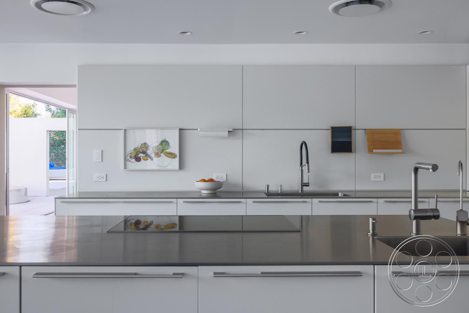 Modern 521 - contemporary art piece on wall, open floor plan with seamless indoor-outdoor flow, interior kitchen with polished concrete countertops, steel kitchen faucet with pull-down sprayer, minimalist cabinetry with flat-panel doors, exposed track lighting fixtures, modern square sink with stainless steel finish, white geometric wall panels, sliding glass doors leading to pool area, light wood accents on kitchen decor, smooth tile backsplash with no grout lines, built-in kitchen shelving for minimalist storage, neutral color palette with accents of green, indoor-outdoor transition with direct view of patio, round fruit bowl as kitchen centerpiece