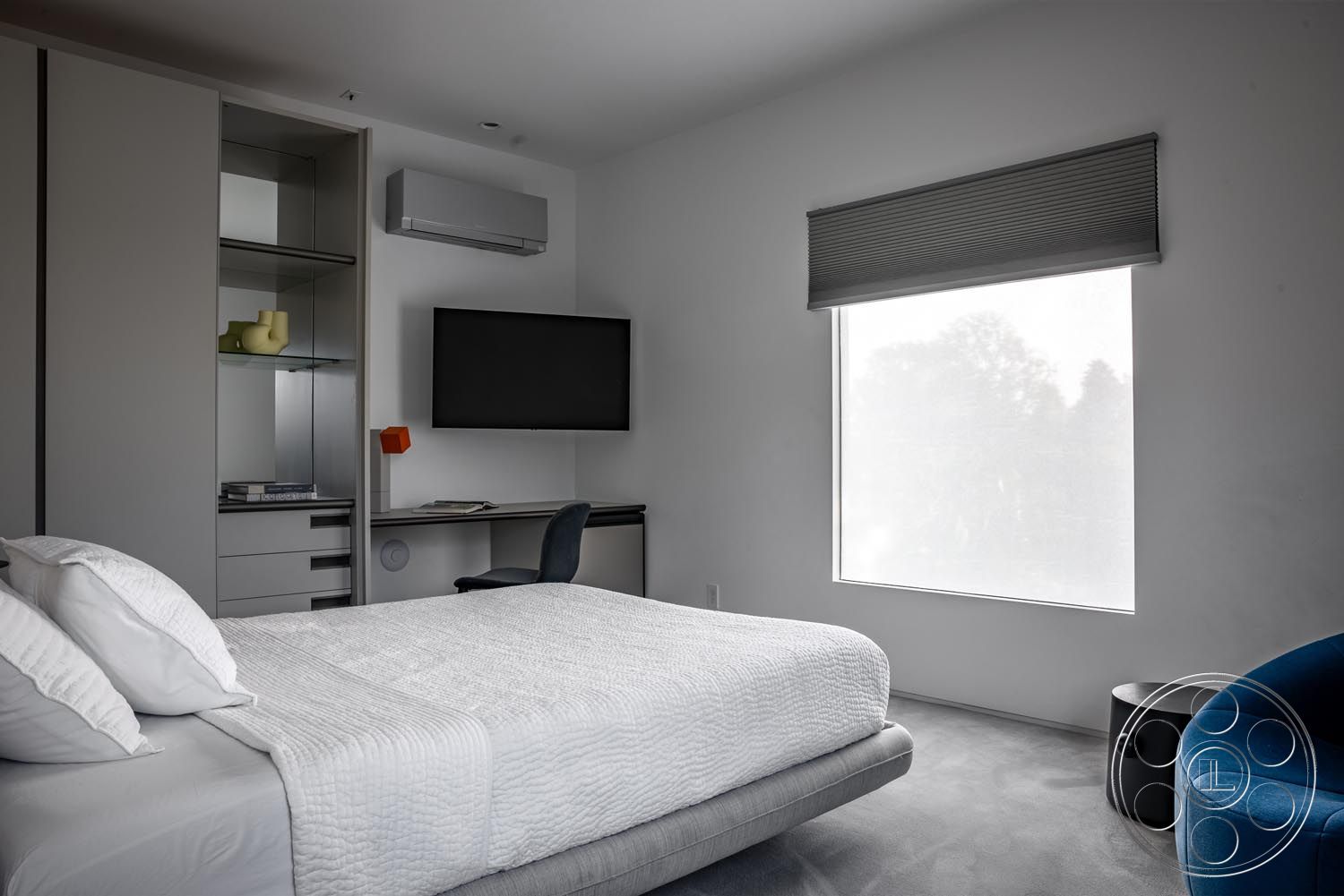Modern 521 - minimalist aesthetic, carpet bedroom flooring, mounted flat-screen television, contemporary desk area with ergonomic chair, modern bedroom interior, calming gray color scheme, king-sized bed with white quilted bedding, clean lines with minimalist design, built-in storage cabinets with glass shelving, floor-to-ceiling window with roller shades, accent chair in blue fabric, subtle abstract decor elements, ambient indoor lighting
