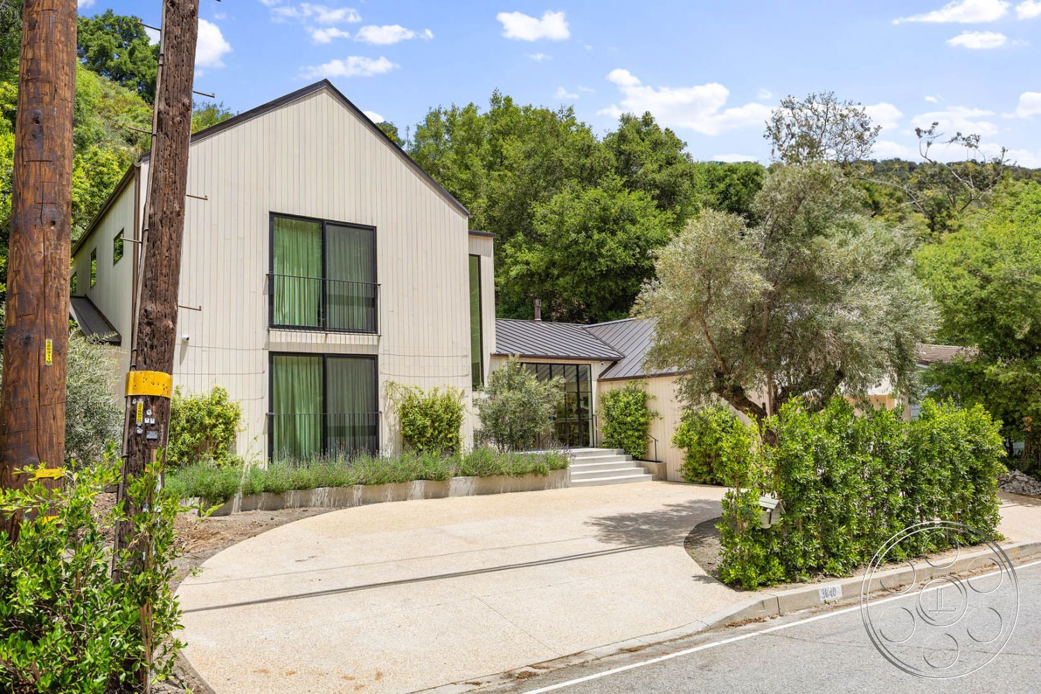 Modern Farmhouse 19 - exterior, modern architectural style, surrounding forest setting, floor-to-ceiling glass windows, sloped hillside location, concrete steps leading to entry, minimalist entryway design, asymmetrical facade design, textured stucco wall cladding, metal roof with angular shape, rectangular driveway with exposed aggregate finish, landscaped front yard with native plants, green tinted glass balcony railings, curved driveway shape, contemporary landscaping elements