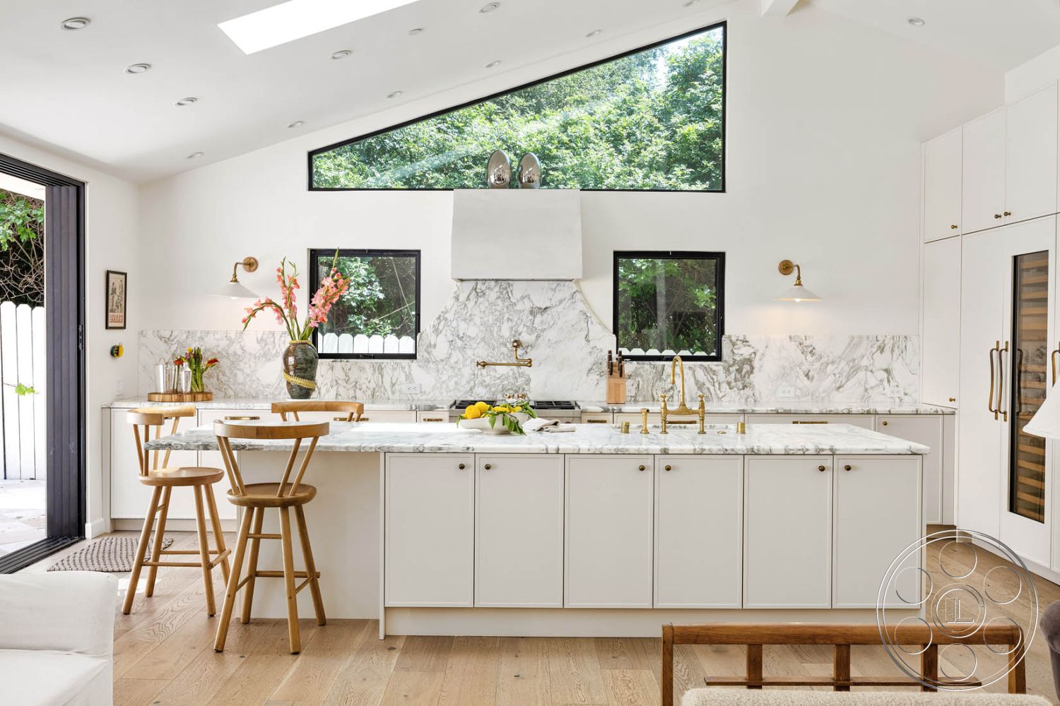 Modern Farmhouse 19 - interior kitchen with marble countertops, minimalistic design with clean lines, white cabinetry with brass hardware, open concept space with vaulted ceiling, wooden bar stools in dining area, large floor-to-ceiling windows providing natural light, textured marble backsplash behind the stove, light hardwood flooring in kitchen, black framed windows for modern contrast, plant decoration on countertop for natural touch, skylights for enhanced lighting, decorative wall art in kitchen area, neutral color palette with light tones, high-efficiency stainless steel appliances, contemporary lighting fixtures over the island