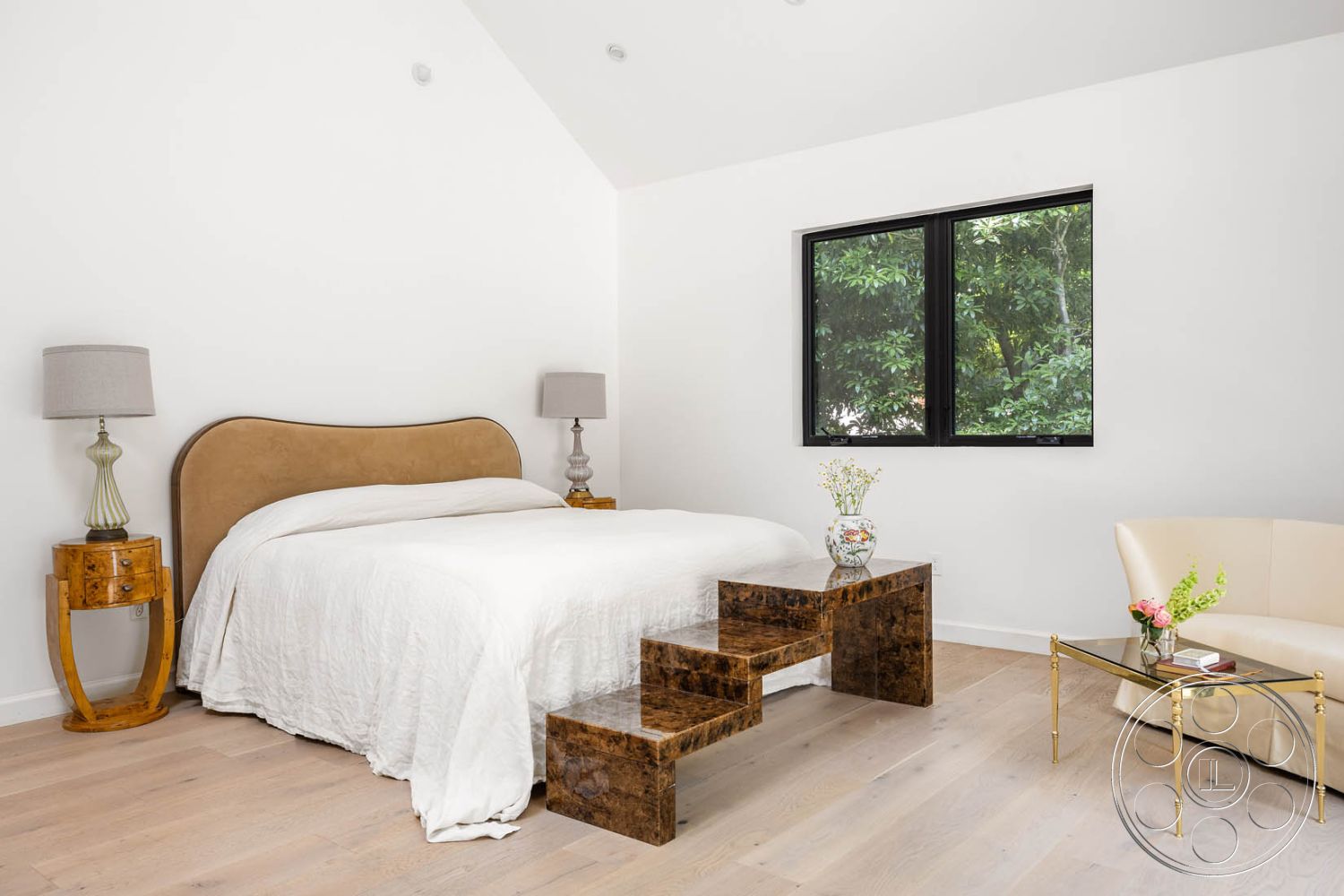 Modern Farmhouse 19 - minimalist aesthetic, vaulted ceiling, black window frames, decorative bedside lamp, warm natural light, hardwood bedroom floors, white painted walls, open and airy layout, mid-century modern armchair, modern bed with curved headboard, glass tabletop bedside table, glass vase with flowers, simple bedding design, side table with unique design