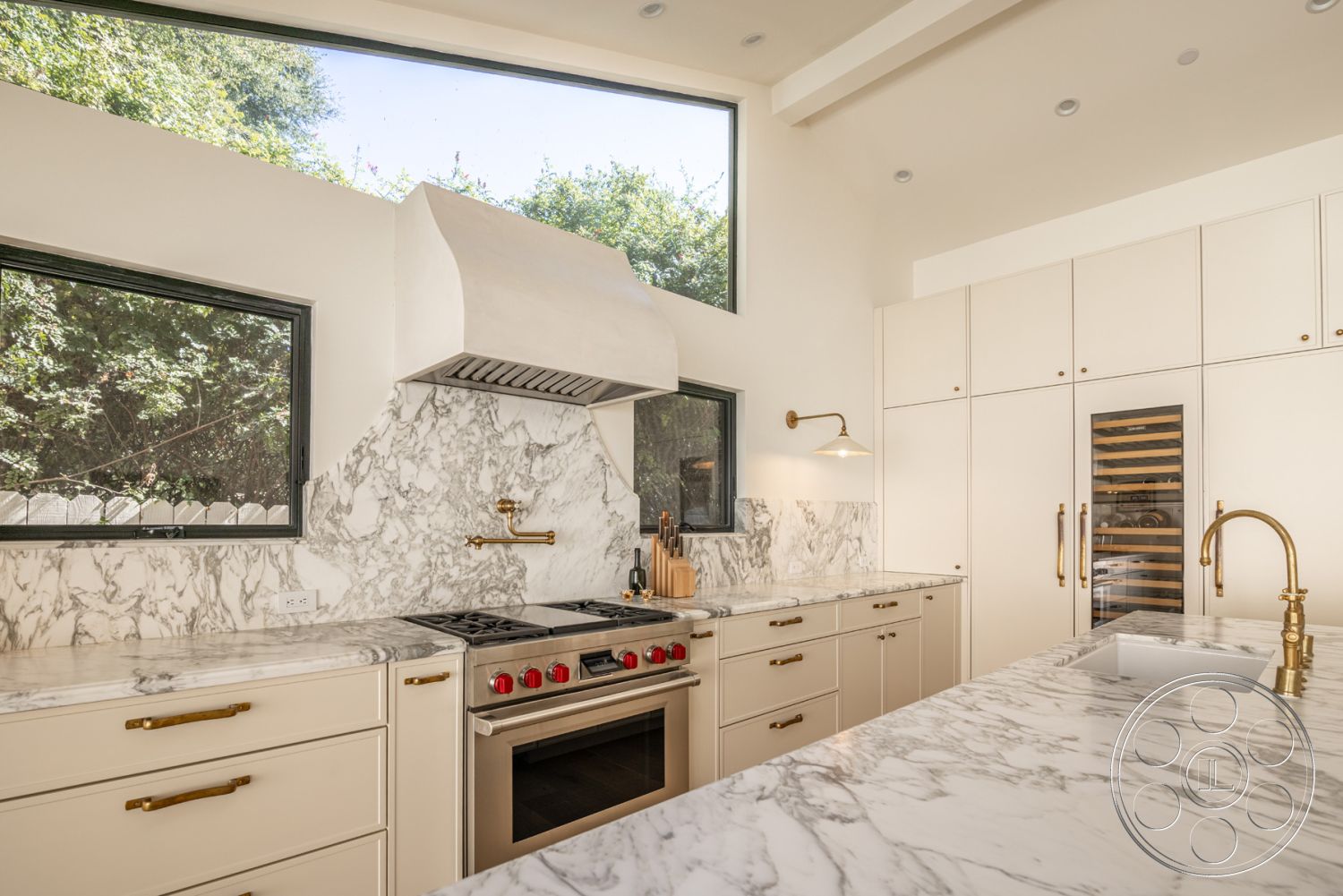 Modern Farmhouse 19 - stainless steel appliances, light-colored cabinetry, contemporary kitchen design, interior kitchen space, marble kitchen countertops, large windows with garden view, marble kitchen backsplash, open floor plan kitchen, high ceilings with recessed lighting, brushed gold faucet fixtures, natural light in kitchen, built-in wine cooler, kitchen island with overhang, exposed hood vent, cabinets with gold hardware