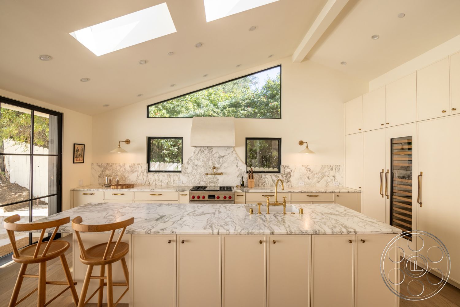 Modern Farmhouse 19 - modern architectural style, gold pendant lighting, black stainless steel appliances, gold faucet fixtures, interior kitchen with marble countertops, textured marble backsplash, hardwood kitchen flooring, skylights for natural lighting, minimalist kitchen layout, integrated wine cooler, white painted kitchen cabinets, marble kitchen island with waterfall edge, concealed refrigerator within cabinetry, open floor plan kitchen design, dark framed windows, subtle cabinetry hardware, angled roof design with high ceilings, outdoor view through large glass doors, comfortable seating with wooden bar stools, natural green surroundings