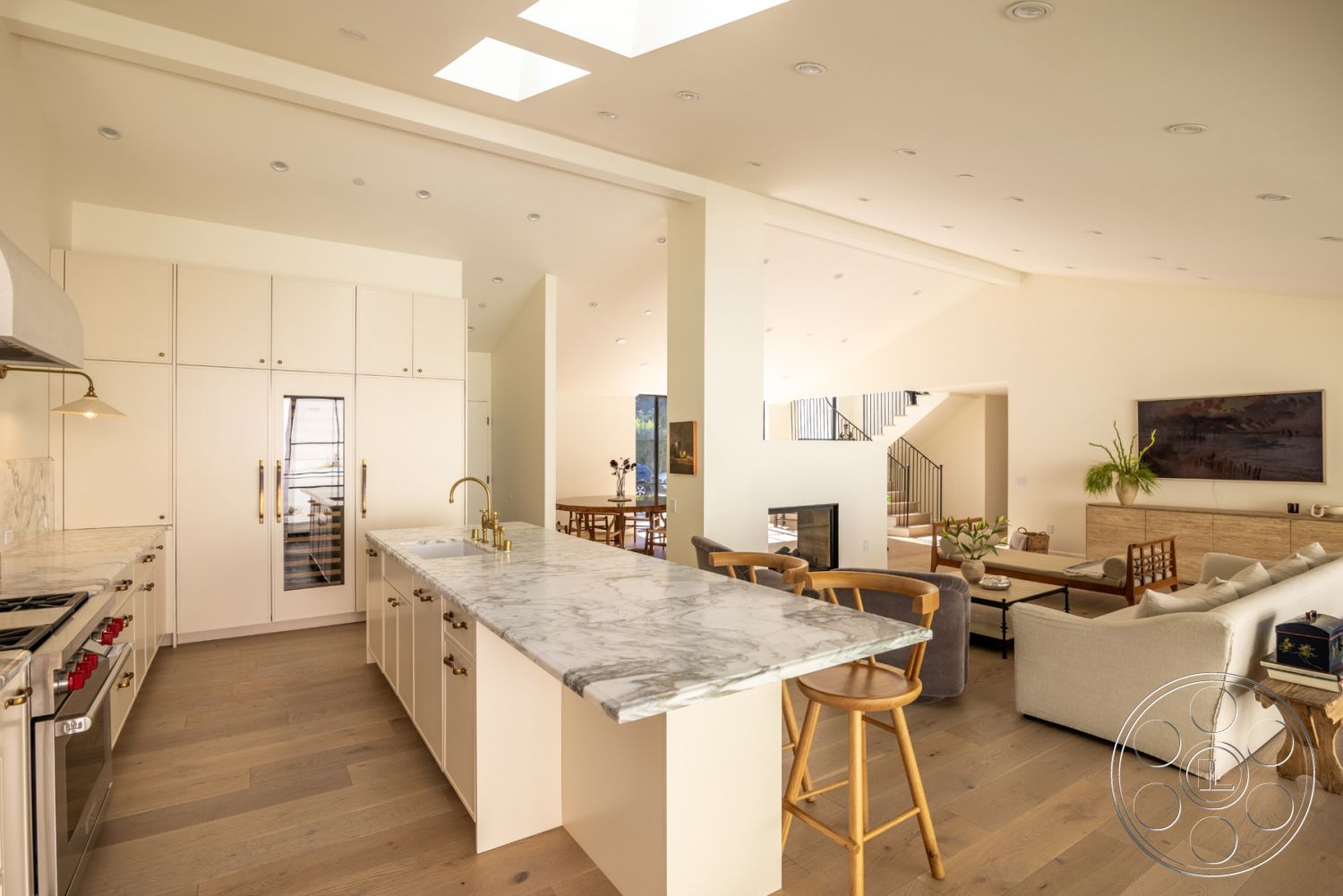 Modern Farmhouse 19 - neutral color palette, large windows for natural light, marble kitchen countertop, minimalist design style, contemporary artwork on walls, skylight ceiling feature, light beige wall paint, open concept interior layout, modern art wall decoration, white cabinetry with brass hardware, hardwood flooring throughout living area, modern pendant lighting above island, exposed metal railing on staircase, contemporary dining area with wooden table, integrated refrigerator in cabinetry, seating area with fabric sofa, built-in storage solutions in living room, wooden bar stools at kitchen island, cozy fireplace in the living area, elevated ceiling height in common area