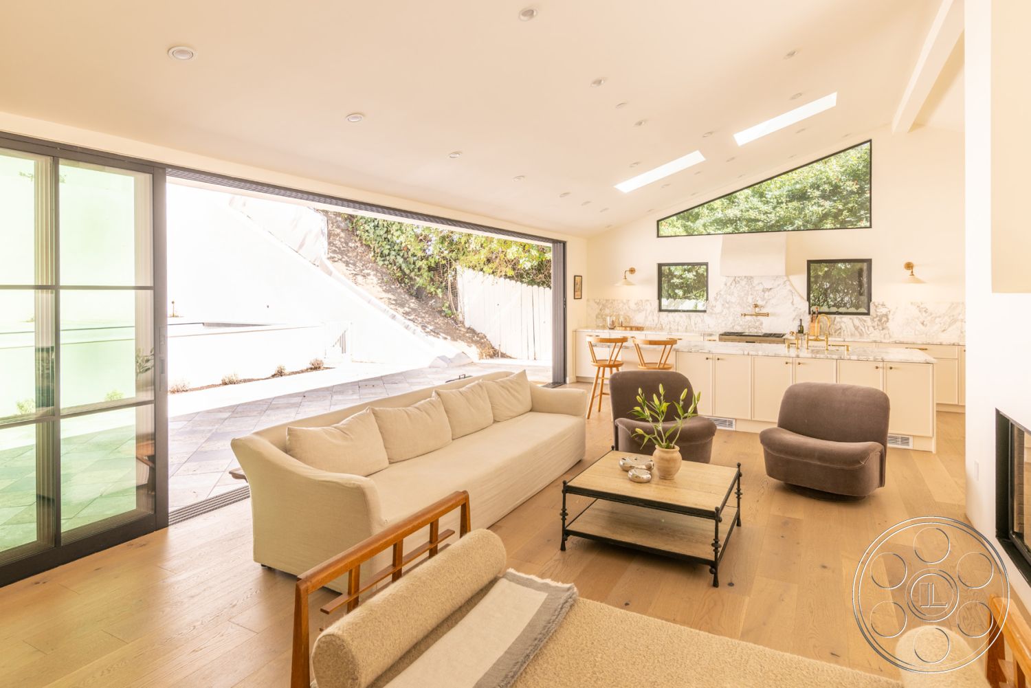 Modern Farmhouse 19 - neutral color palette, large glass sliding doors, modern light fixtures, custom cabinetry, black window frames, textured wall finishes, indoor-outdoor living, light wood dining table, exposed ceiling beams, hardwood living room floors, natural light from skylights, exterior landscaped garden view, minimalist furniture design, open concept interior, contemporary wall decor, spacious and airy layout, angled roof design, modern kitchen with marble countertops, sloped backyard terrain, lush greenery visible outside