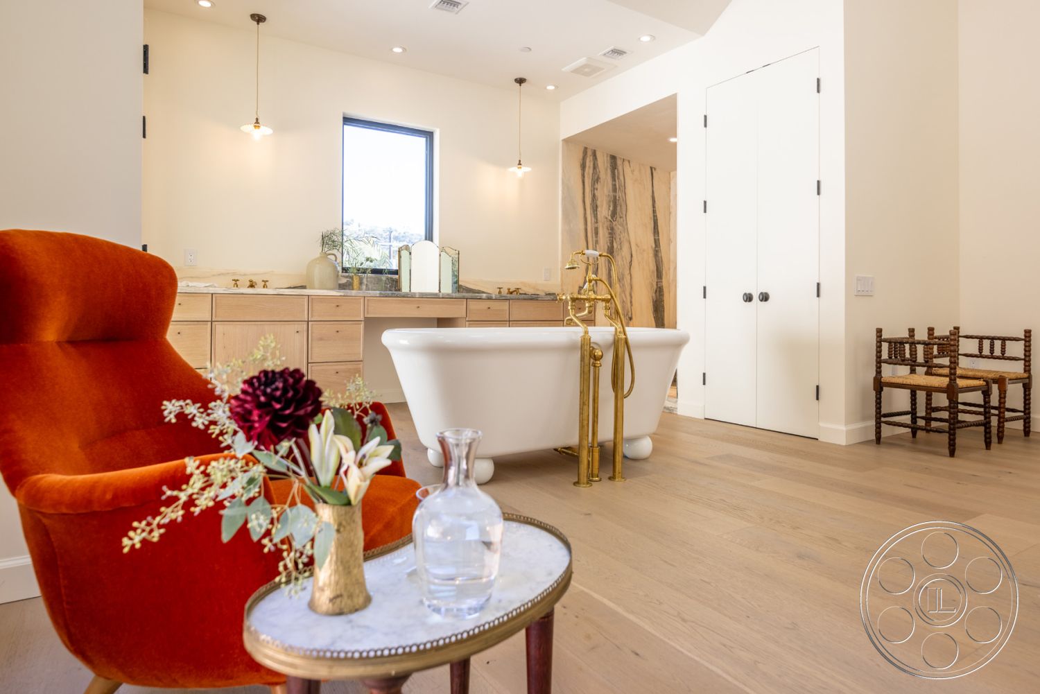 Modern Farmhouse 19 - neutral color palette, mid-century modern accent chair, minimalist pendant lighting, white painted drywall, large window with modern trim, interior hardwood bathroom flooring, freestanding porcelain bathtub, golden brass tub faucet, wooden vanity cabinets with natural finish, textured stone bathroom wall, decorative glass vase with flowers, satin brass cabinet hardware, minimalist round accent table, white double doors with black knobs, small rustic wooden stool