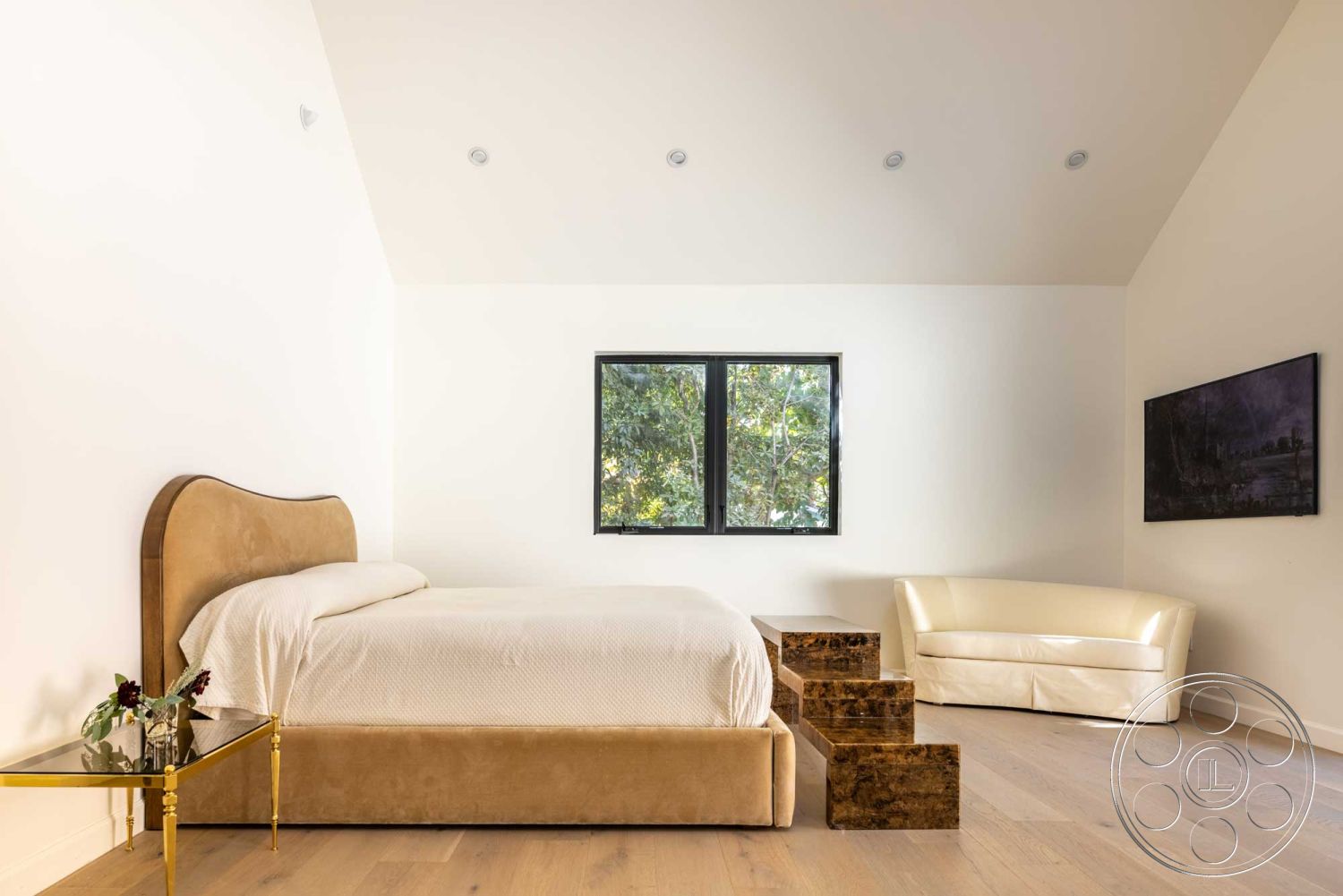 Modern Farmhouse 19 - open space layout, high vaulted ceiling, natural light from window, neutral toned bedding, contemporary art on wall, interior bedroom with wood plank flooring, upholstered headboard bed, floor-to-ceiling window with black frame, minimalist decor with modern furniture, gold metal bedside table, textured light-colored walls, stone steps leading to bed, curved sofa with faux leather upholstery, plant accents near window, calm and serene color palette