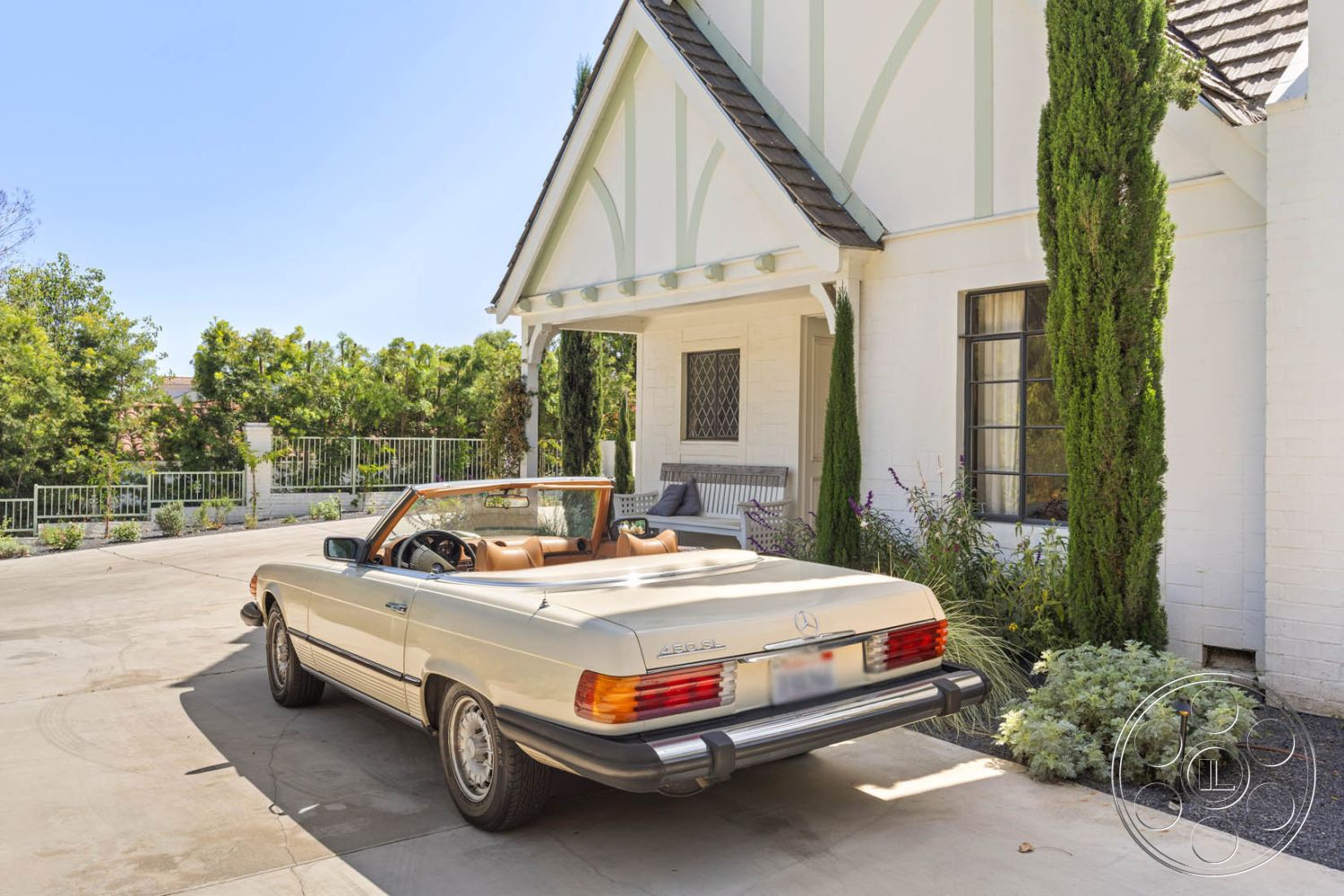 English 60 - wooden bench seating, tall evergreen trees, urban residential setting, Mediterranean architectural style, gabled roof with asphalt shingles, exterior white brick walls, metal window frames, grey asphalt driveway, landscaped garden with low shrubs, front porch with decorative wood supports, natural stone accent pathways, adjacent green lawn area, cobblestone border around garden beds