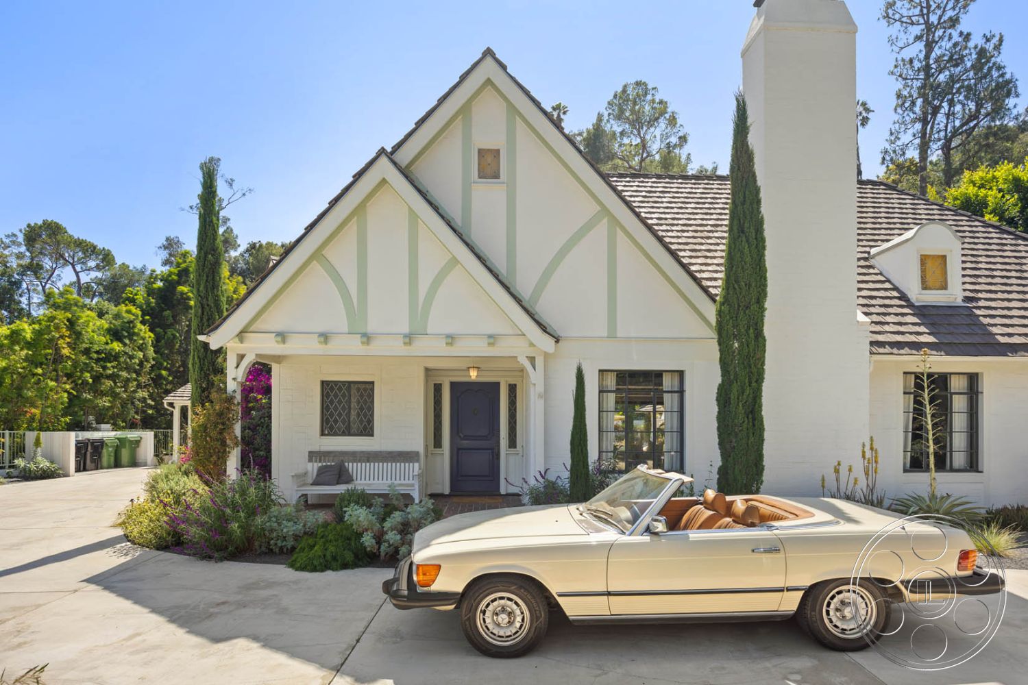 English 60 - concrete driveway, arched doorways, exposed brick chimney, slate roof shingles, exterior white painted walls, surrounded by lush greenery, craftsman style front porch, vaulted entryway ceiling, landscaped garden patio with flowers, tall cypress trees flanking entrance, decorative gable ends with wood accents, stone pathway leading to front door, large double-hung windows with grid pattern, single-car vintage convertible parked in driveway, sunny residential neighborhood setting