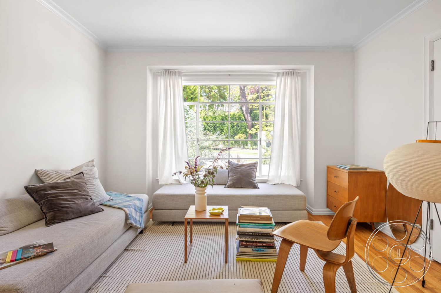 English 60 - light hardwood flooring, soft natural light, decorative throw pillows, view of greenery outside, interior living room, natural fiber area rug, large picture window, white painted walls, potted indoor plants, minimalist sofa with gray upholstery, mid-century modern wooden coffee table, floor lamp with paper shade, modern wooden sideboard