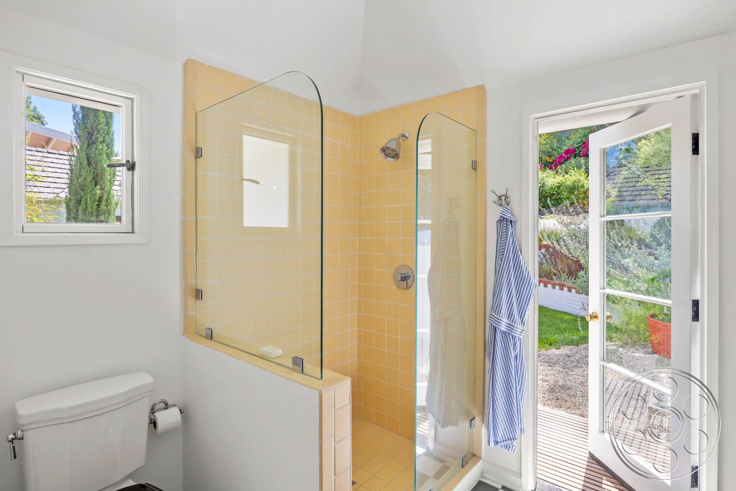 English 60 - natural light, textured flooring, minimalist aesthetic, white porcelain toilet, outdoor garden view, glass shower doors, interior bathroom, modern bathroom design, white painted walls, stone patio flooring, wooden door with glass panels, yellow ceramic tile shower walls, framed window, towel hanging hook, shower head fixture