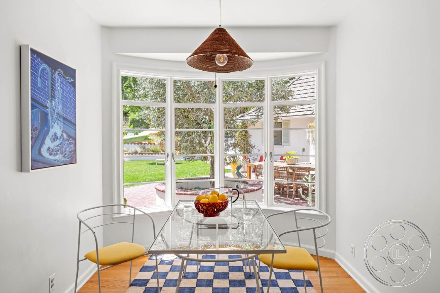 English 60 - lush greenery, glass dining table, clear acrylic chairs, decorative fruit bowl, woven pendant light fixture, wooden floorboards, white painted walls, curved window design, large bay windows, view of outdoor patio, interior dining nook, yellow upholstered seat cushions, checkered blue and white area rug, brick patio area