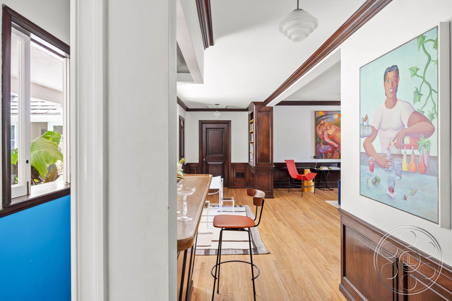 English 60 - interior dark wood paneling, interior room with natural light, interior coffered ceiling details, interior hardwood living room floors, interior white painted walls, interior open floor plan, interior crown molding details, interior modern art wall decor, interior circular bar table, interior sleek barstool design, interior decorative glass light fixture, interior built-in shelving in hallway, interior framed paintings on walls, interior color accent wall with blue paint, interior stylish dining area with colorful chairs