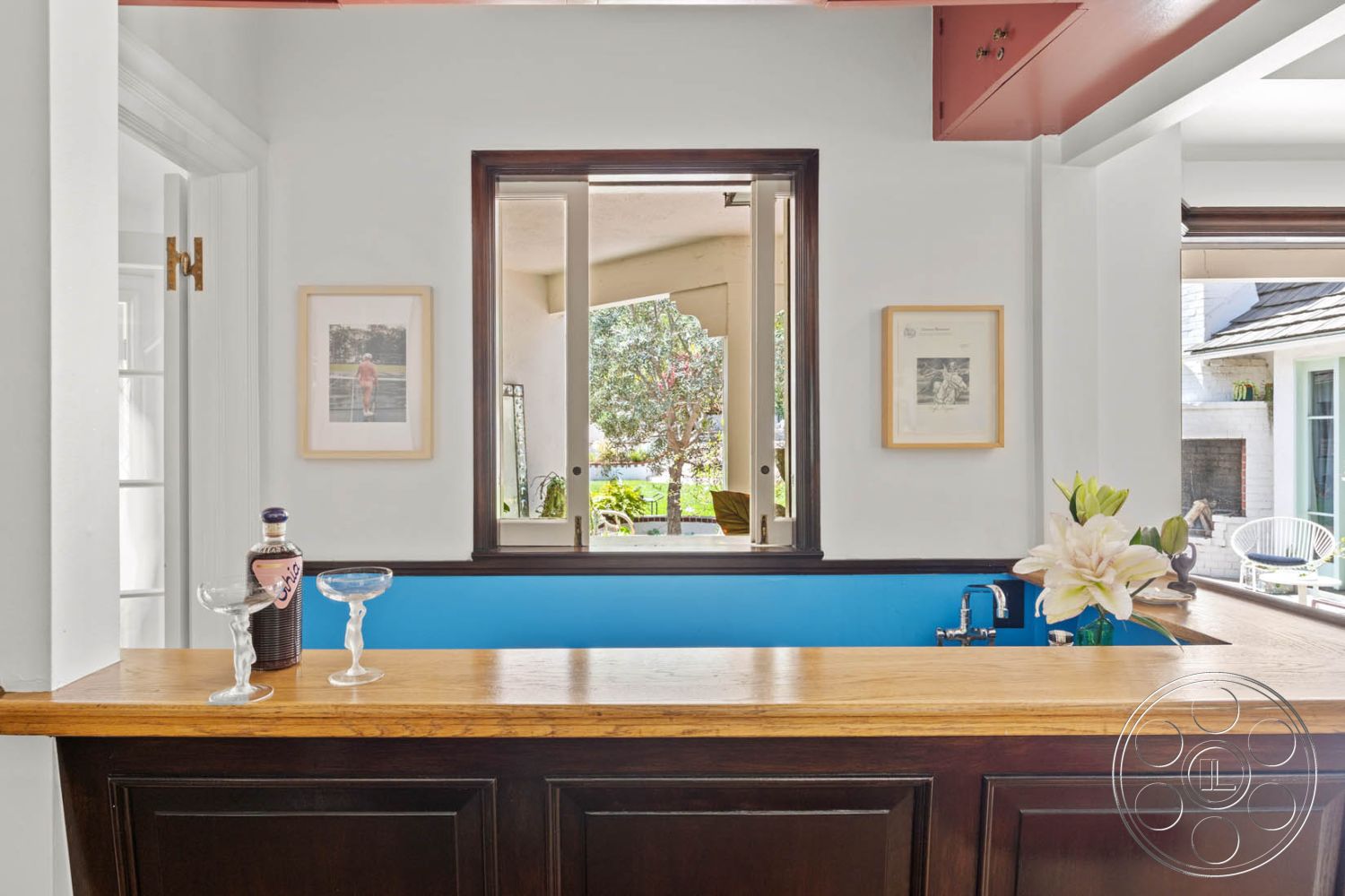 English 60 - dark wood cabinetry, casual entertaining space, white painted walls, open floor plan design, natural light-filled room, exterior garden view, decorative flower arrangement, stainless steel bar sink, interior wooden countertop bar, natural wood bar top, large window framing garden, modern framed artwork, blue accent wall behind counter, floating bar design