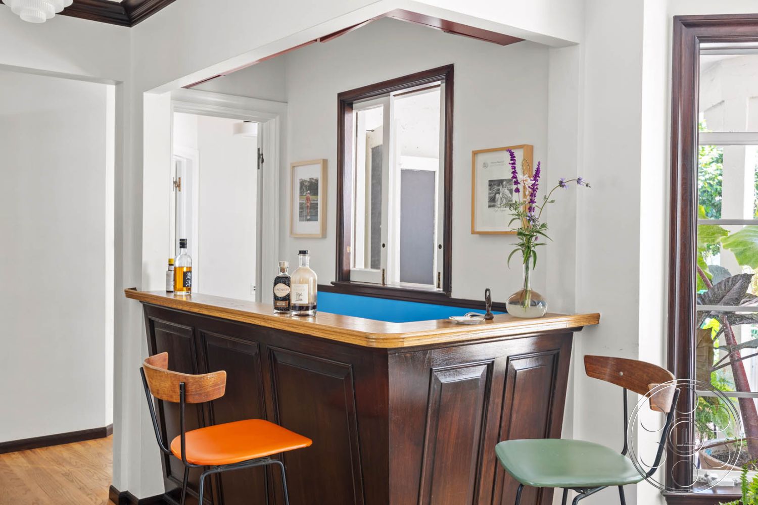 English 60 - modern minimalist decor, hardwood flooring in living area, interior bar countertop with wooden surface, exposed wood bar front with paneling, dark wood trim around windows, open layout kitchen and bar area, light-colored walls with white paint, simple glass window panes, decorative flower vase on bar, colorful bar stool seating in orange and green, natural light from windows creating bright ambiance, artwork framed on wall adjacent to bar, contemporary home interior design, stylish home bar setup