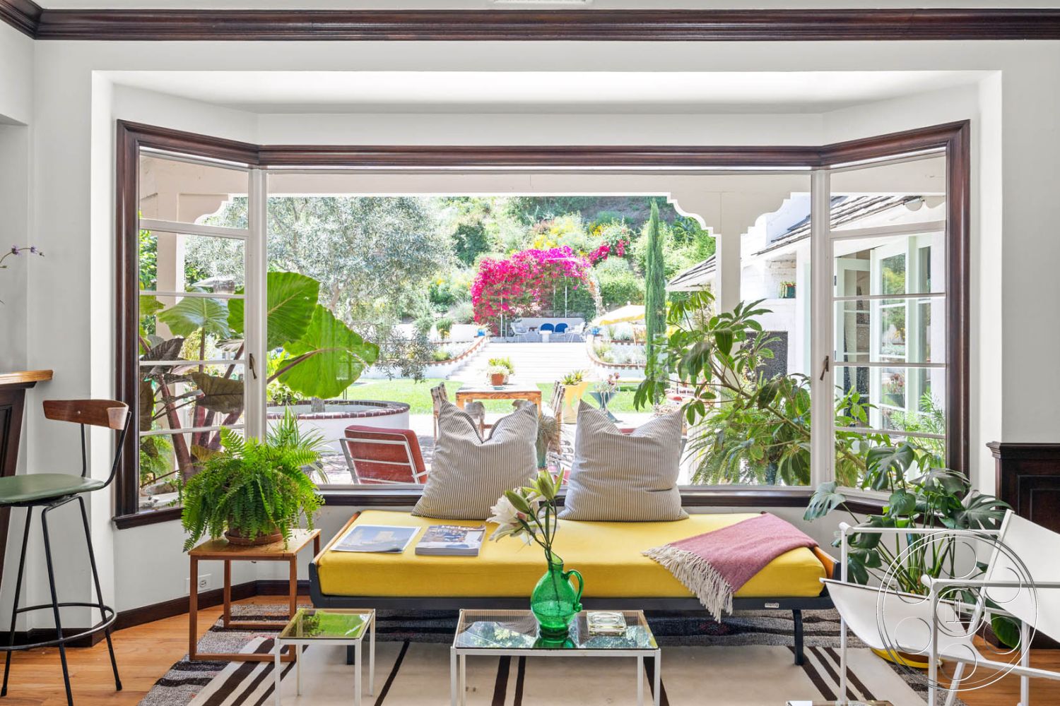 English 60 - glass coffee table, open floor plan, cozy reading nook, landscaped garden view, white painted walls, potted indoor plants, hardwood flooring in living room, natural light from large windows, contemporary interior decor, exposed wood trim, bougainvillea plants, bay window design, modern yellow sofa, dark wood barstool, black and white area rug, open view to exterior patio, circular patio