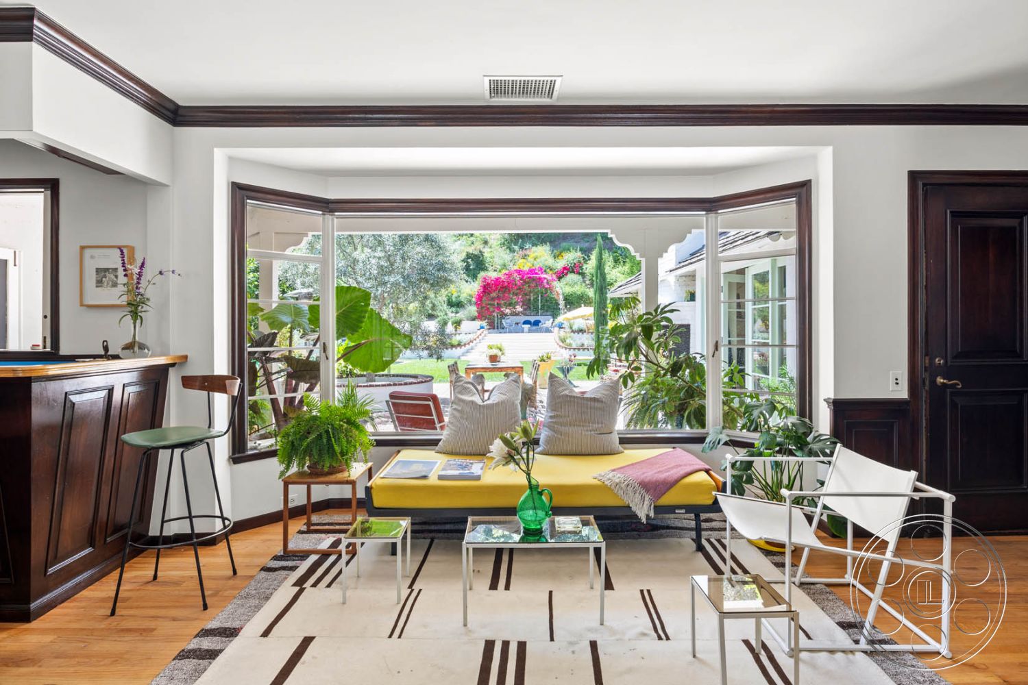 English 60 - natural light-filled space, hardwood floor living room, white painted walls interior, modern minimalist furniture, urban residential environment, inviting indoor-outdoor flow, exposed wood trim around windows, built-in bar with dark wood cabinetry, floor-to-ceiling windows with garden view, geometric area rug with stripes, contemporary glass coffee tables, indoor plants adding greenery, cushioned seating with throw pillows, stainless steel barstool with green seat, black wood accent wall, lush garden patio setting, colorful blooming flowers visible outside, relaxed casual lounge area