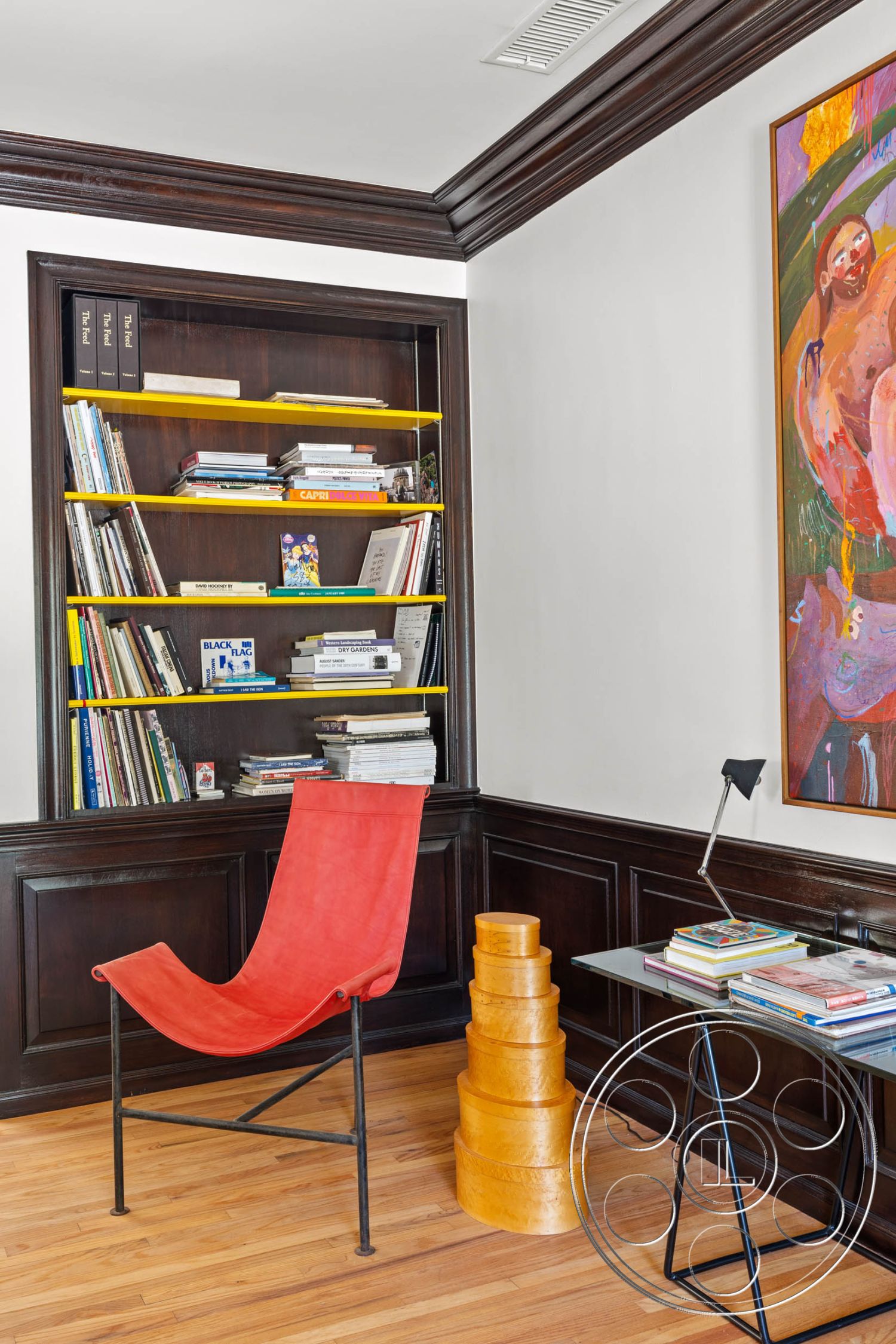 English 60 - minimalist design, hardwood floor, dark wood paneling, modern bookshelf, white painted walls, crown molding details, glass side table, colorful abstract wall art, contemporary style room, yellow accent shelves, red fabric chair
