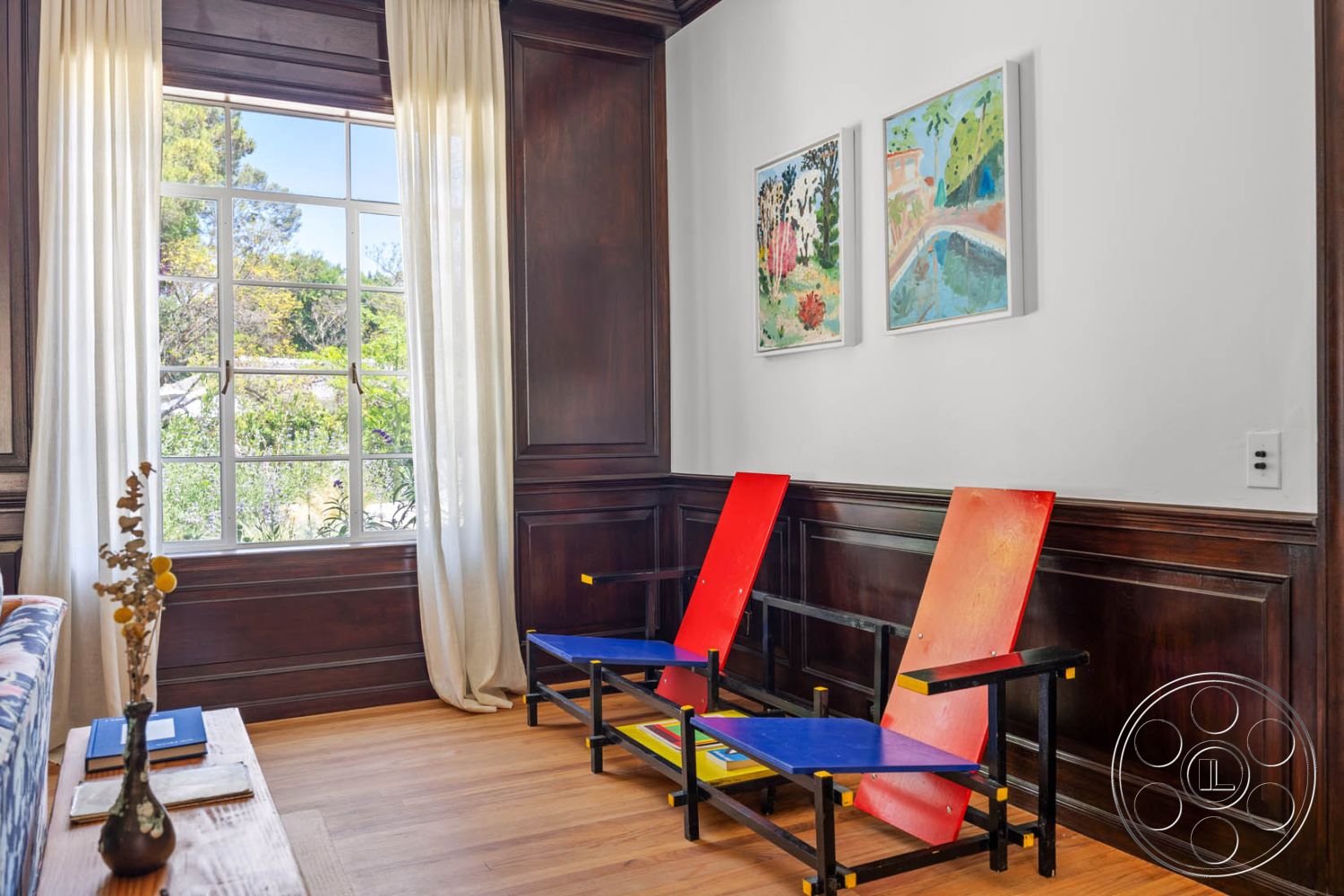 English 60 - cozy reading nook, hardwood living room floors, modern wooden coffee table, dark wood paneling interior, white painted walls interior, natural light-filled sitting area, floor-to-ceiling window with garden view, double-hung window with grid pattern, decorative wall art in frame, light curtains with soft drape, colorful contemporary chairs, exposed wood trim around window, suburban interior setting